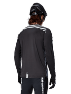 Alpinestars A-Dura Dri Oscar långärmad tröja, Black Reflective, vy bakifrån som visar reflekterande diagonala ränder på axlarna och ett förlängt ryggslut, buren med svart MTB-hjälm och handskar, teknisk utrustning för mountainbike