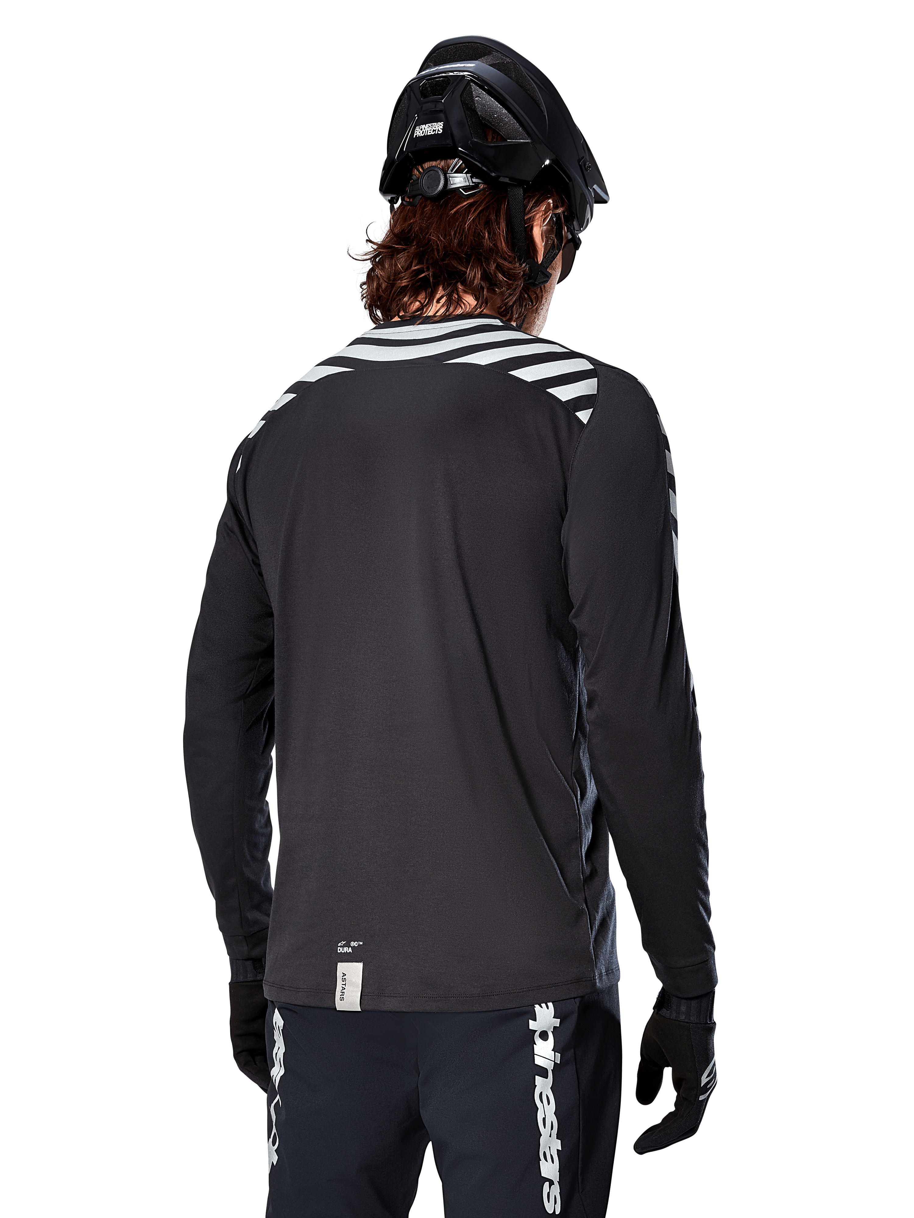Alpinestars A-Dura Dri Oscar långärmad tröja, Black Reflective, vy bakifrån som visar reflekterande diagonala ränder på axlarna och ett förlängt ryggslut, buren med svart MTB-hjälm och handskar, teknisk utrustning för mountainbike