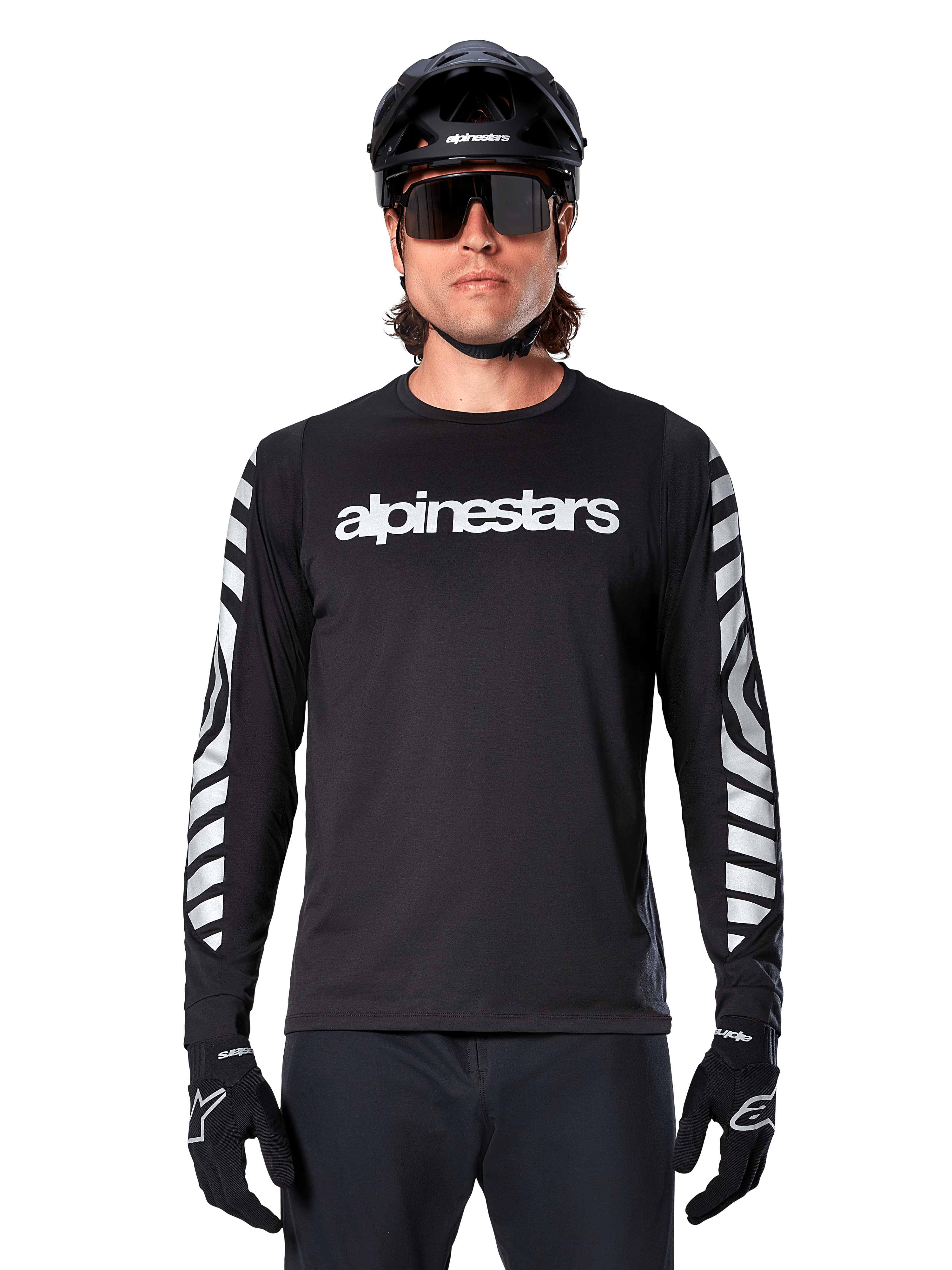 Manlig cyklist i Alpinestars A-Dura Dri Oscar långärmad tröja, Black Reflective, med vit Alpinestars-text på bröstet och reflekterande silvergrafik på ärmarna, kombinerat med svart MTB-hjälm, solglasögon och svarta handskar, teknisk cykelutrustning