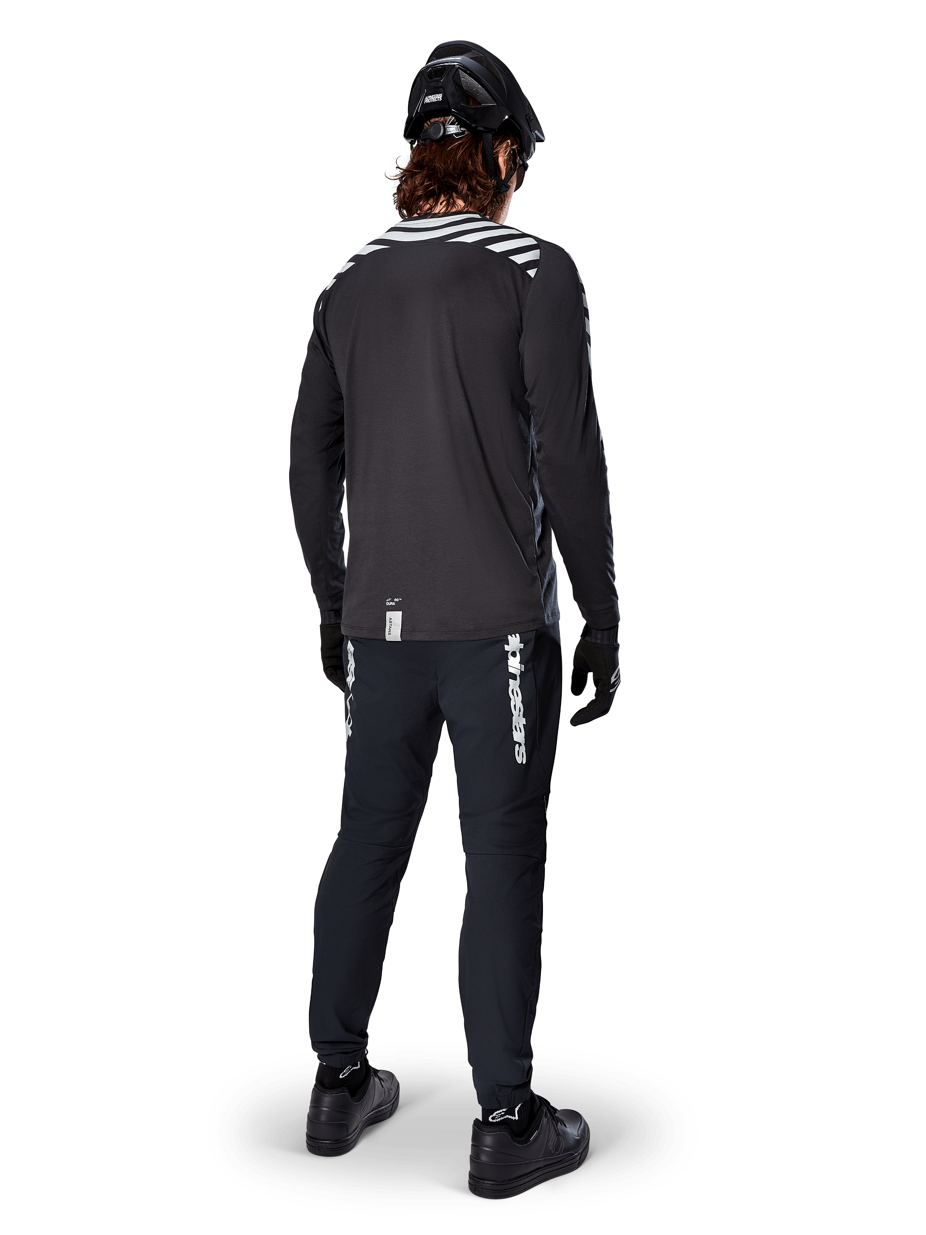Mountainbike-cyklist som bär Alpinestars A-Dura Dri Oscar långärmad tröja i Black Reflective, sedd bakifrån, kombinerad med svarta tekniska byxor, hjälm och handskar, med reflekterande ränder på axlarna och ett förlängt ryggslut för terrängcykling.