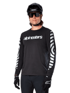 Alpinestars A-Dura Dri Oscar långärmad tröja, MTB-tröja, svart med reflekterande detaljer, vit textlogotyp på bröstet och framträdande reflekterande cirkulär grafik på ärmarna, bärs med svart hjälm och solglasögon, tekniskt fukttransporterande material för terrängcykling