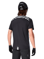 Alpinestars A-Dura Dri Oscar-tröja, kortärmad MTB-tröja, Black Reflective, ryggvy på modell som bär cykelhjälm och handskar, med reflekterande grå axelränder och förlängt ryggslut, fukttransporterande tekniskt material för mountainbike
