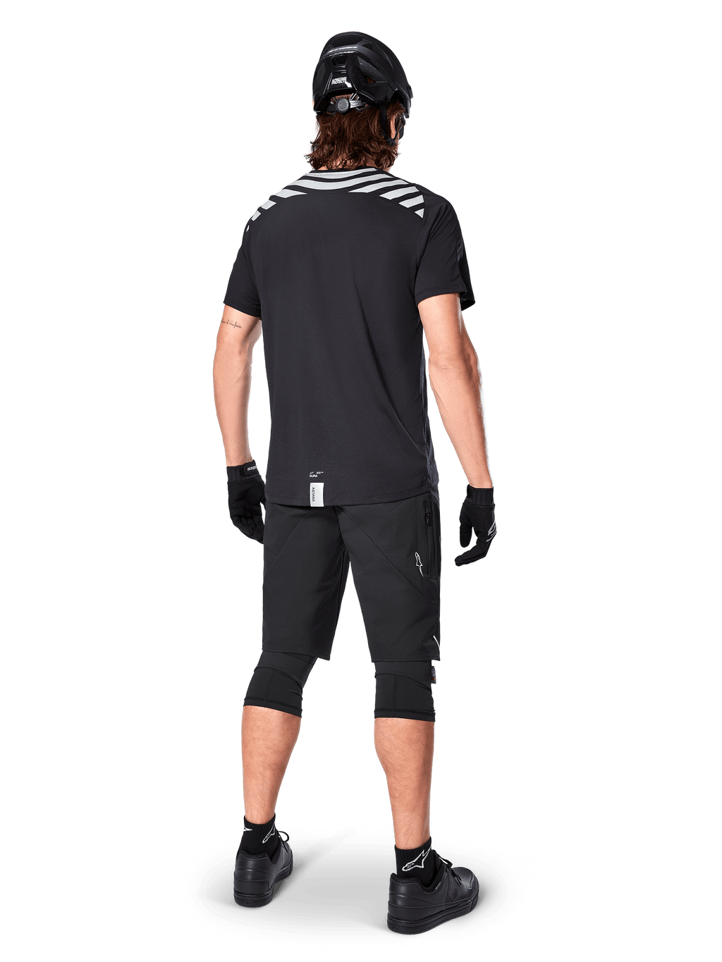 Alpinestars A-Dura Dri Oscar kortärmad tröja, Black Reflective, vy bakifrån av en cyklist som bär tröjan med svarta mountainbikeshorts, hjälm och knäskydd, med reflekterande grafik på axlarna och förlängt ryggslut för MTB-cykling.