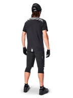 Alpinestars A-Dura Dri Oscar kortärmad tröja, Black Reflective, vy bakifrån av en cyklist som bär tröjan med svarta mountainbikeshorts, hjälm och knäskydd, med reflekterande grafik på axlarna och förlängt ryggslut för MTB-cykling.