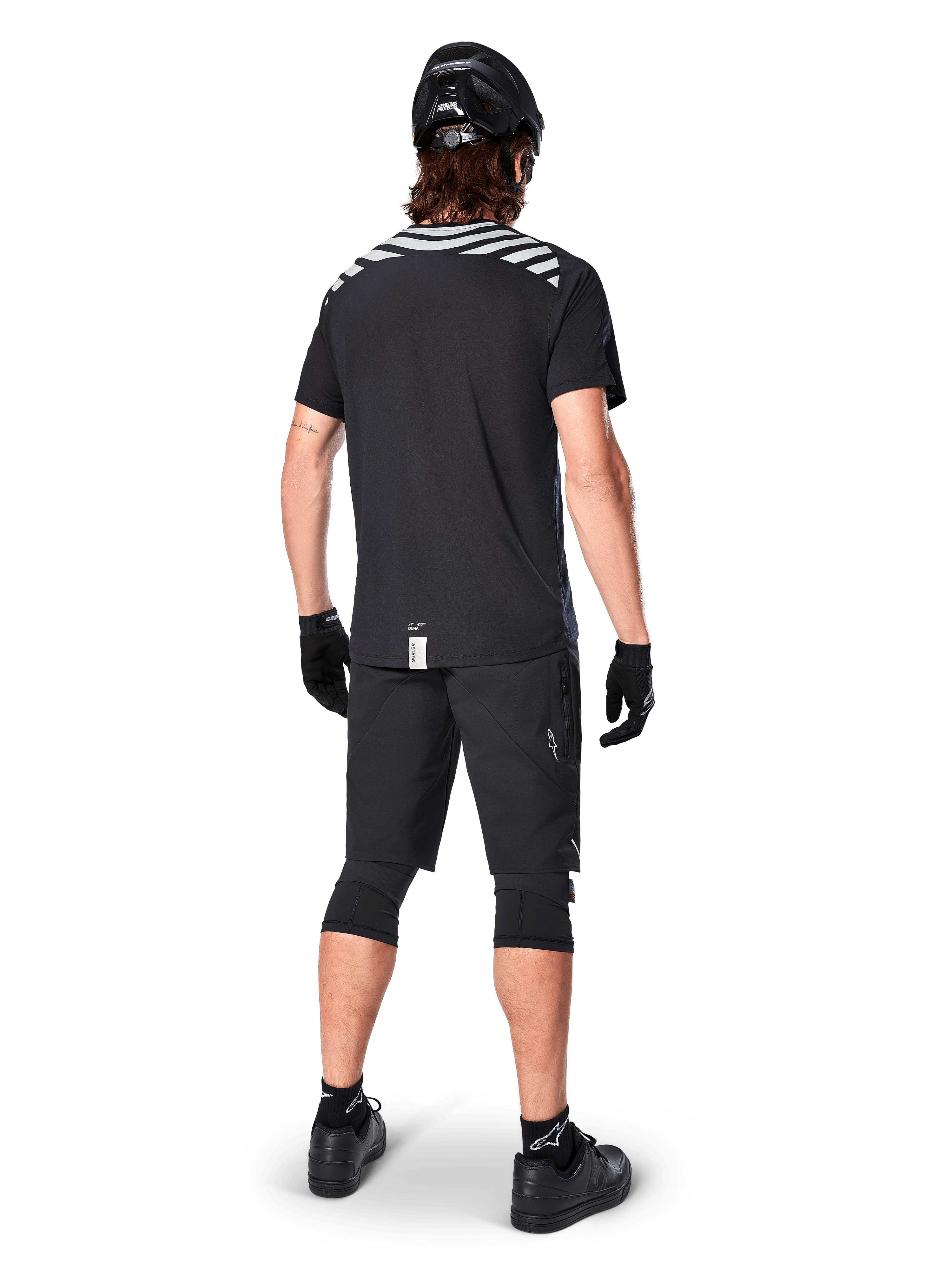 Alpinestars A-Dura Dri Oscar kortärmad tröja, Black Reflective, vy bakifrån av en cyklist som bär tröjan med svarta mountainbikeshorts, hjälm och knäskydd, med reflekterande grafik på axlarna och förlängt ryggslut för MTB-cykling.