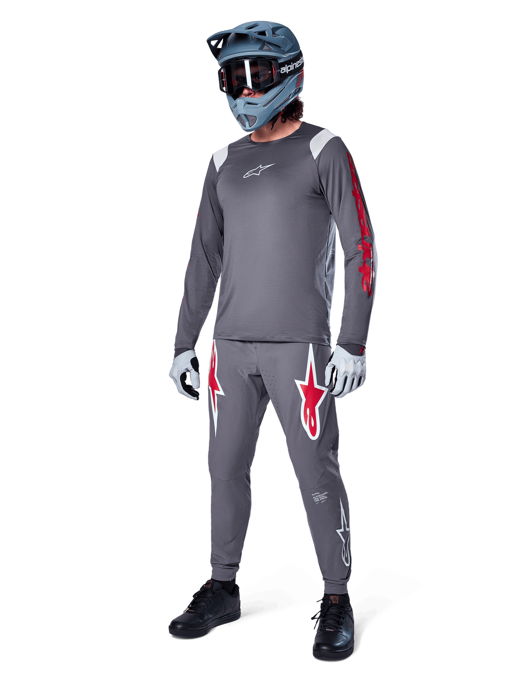 Alpinestars A-Supra Astar långärmad tröja i mörkgrått, med röd grafik på ärmarna och en vit logotyp på bröstet, buren av en cyklist med matchande gråa byxor, en blågrå fullfacehjälm och tekniska handskar, koordinerad mountainbike-utrustning för gravity-cykling