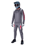Alpinestars A-Supra Astar långärmad tröja i mörkgrått, med röd grafik på ärmarna och en vit logotyp på bröstet, buren av en cyklist med matchande gråa byxor, en blågrå fullfacehjälm och tekniska handskar, koordinerad mountainbike-utrustning för gravity-cykling