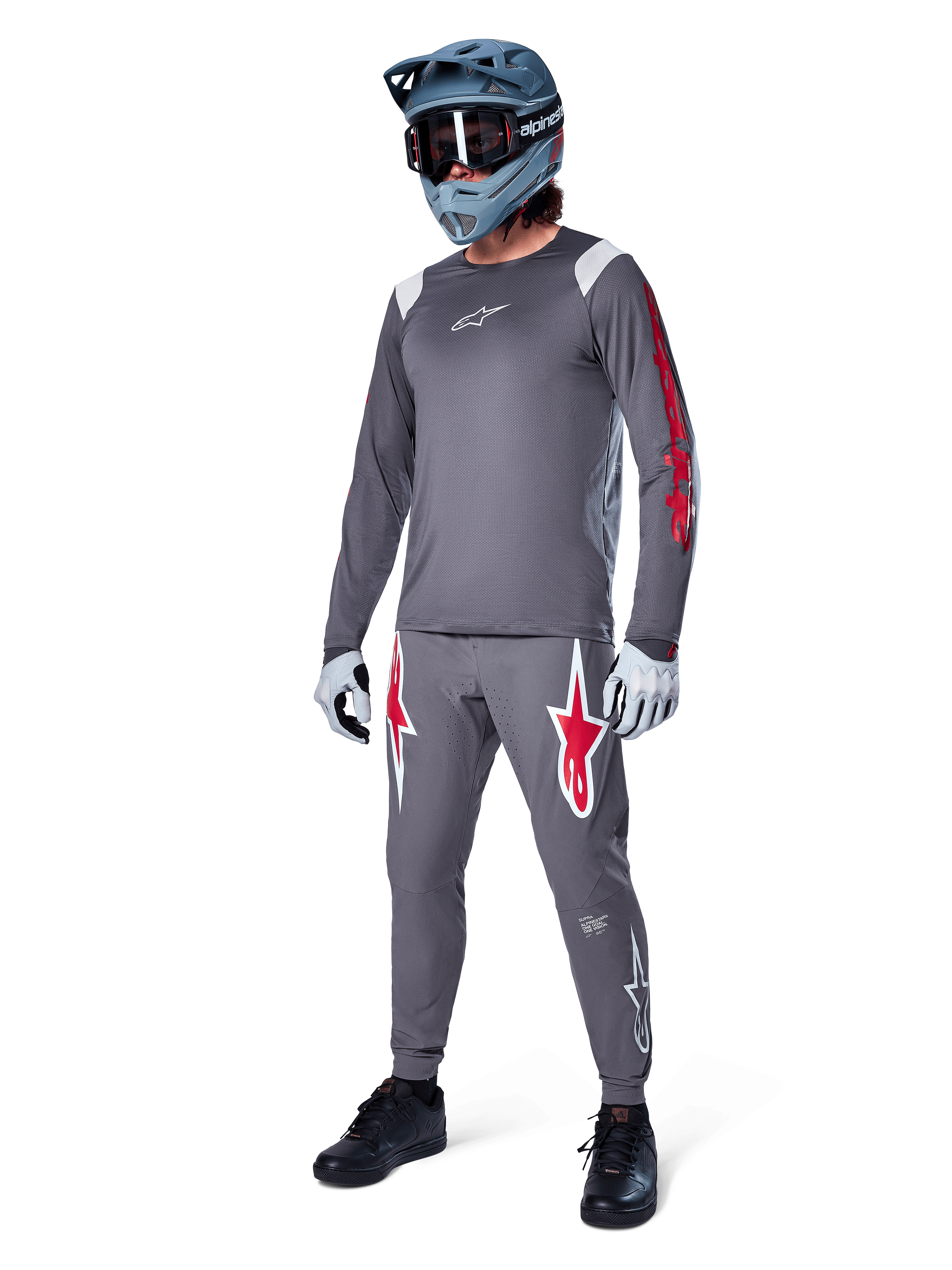 Alpinestars A-Supra Astar långärmad tröja i mörkgrått, med röd grafik på ärmarna och en vit logotyp på bröstet, buren av en cyklist med matchande gråa byxor, en blågrå fullfacehjälm och tekniska handskar, koordinerad mountainbike-utrustning för gravity-cykling