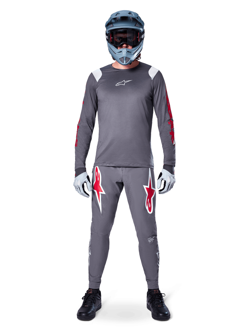 MTB-cyklist i Alpinestars A-Supra Astar långärmad tröja, mörkgrå med röda detaljer och vit bröstlogotyp, matchande grå byxor med stora röda stjärngrafiker, blå fullfacehjälm med goggles, ljusgrå handskar och svarta skor, professionell mountainbike-utrustning