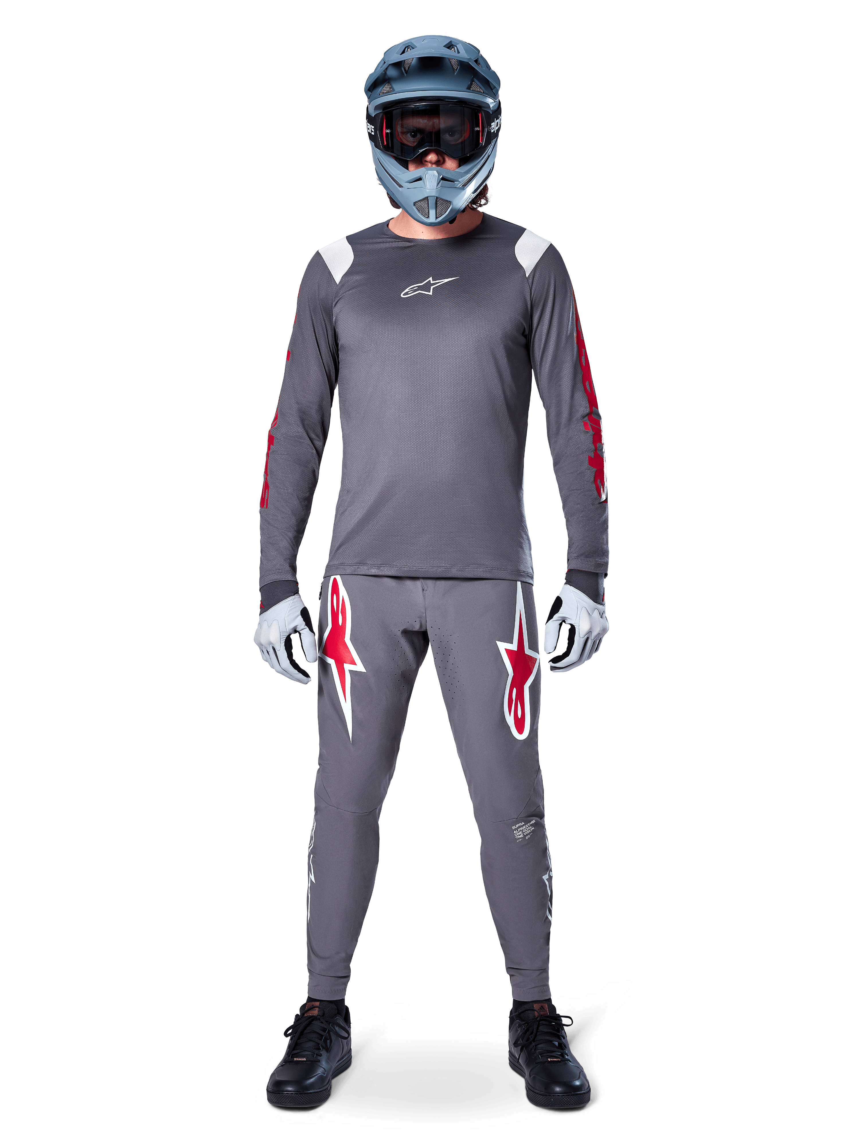 MTB-cyklist i Alpinestars A-Supra Astar långärmad tröja, mörkgrå med röda detaljer och vit bröstlogotyp, matchande grå byxor med stora röda stjärngrafiker, blå fullfacehjälm med goggles, ljusgrå handskar och svarta skor, professionell mountainbike-utrustning