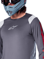 Alpinestars A-Supra Astar långärmad tröja, mörkgrå med vita axelpaneler och röd grafik på ärmarna, bärs med en ljus blågrå fullfacehjälm, funktionskläder för mountainbike