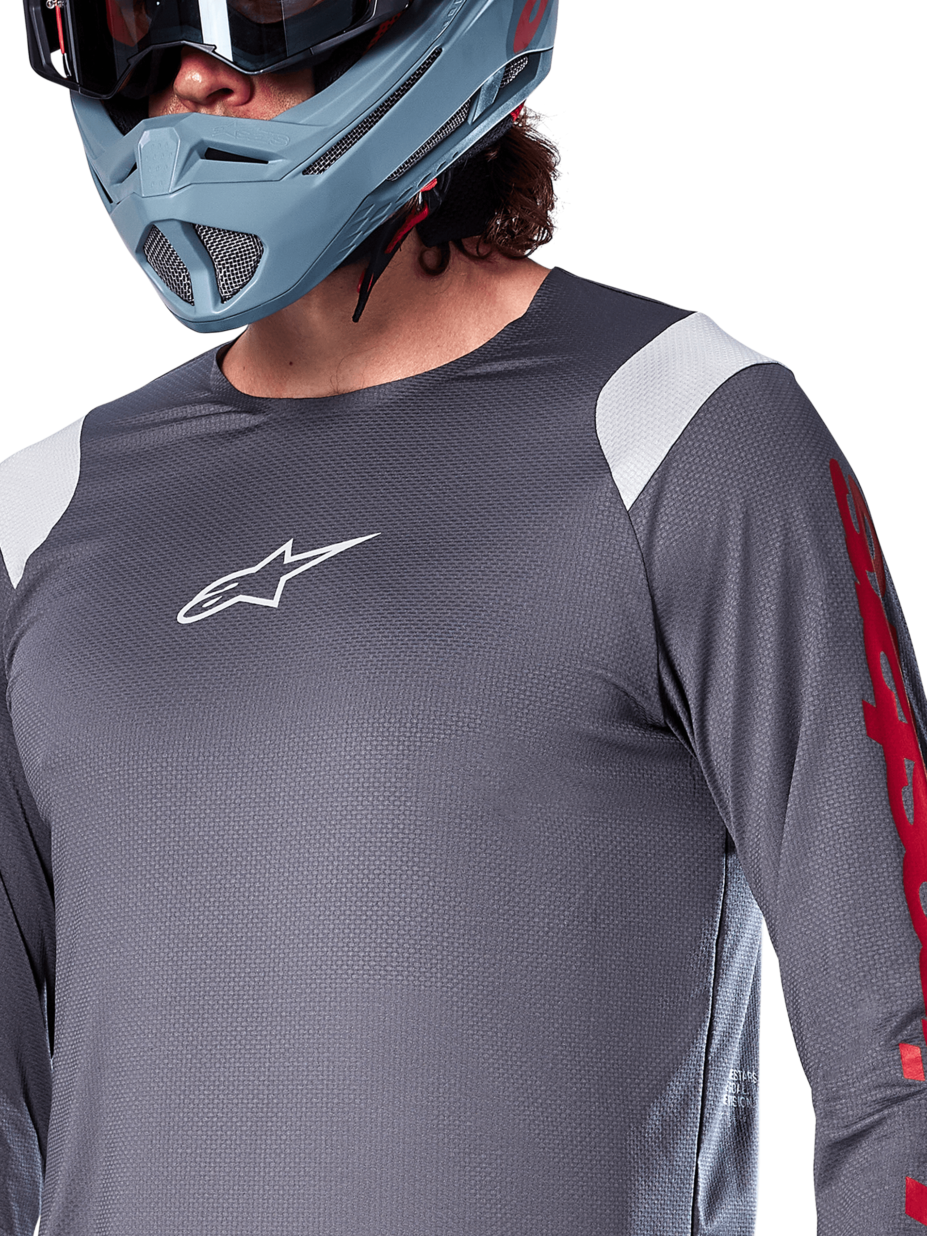 Alpinestars A-Supra Astar långärmad tröja, mörkgrå med vita axelpaneler och röd grafik på ärmarna, bärs med en ljus blågrå fullfacehjälm, funktionskläder för mountainbike