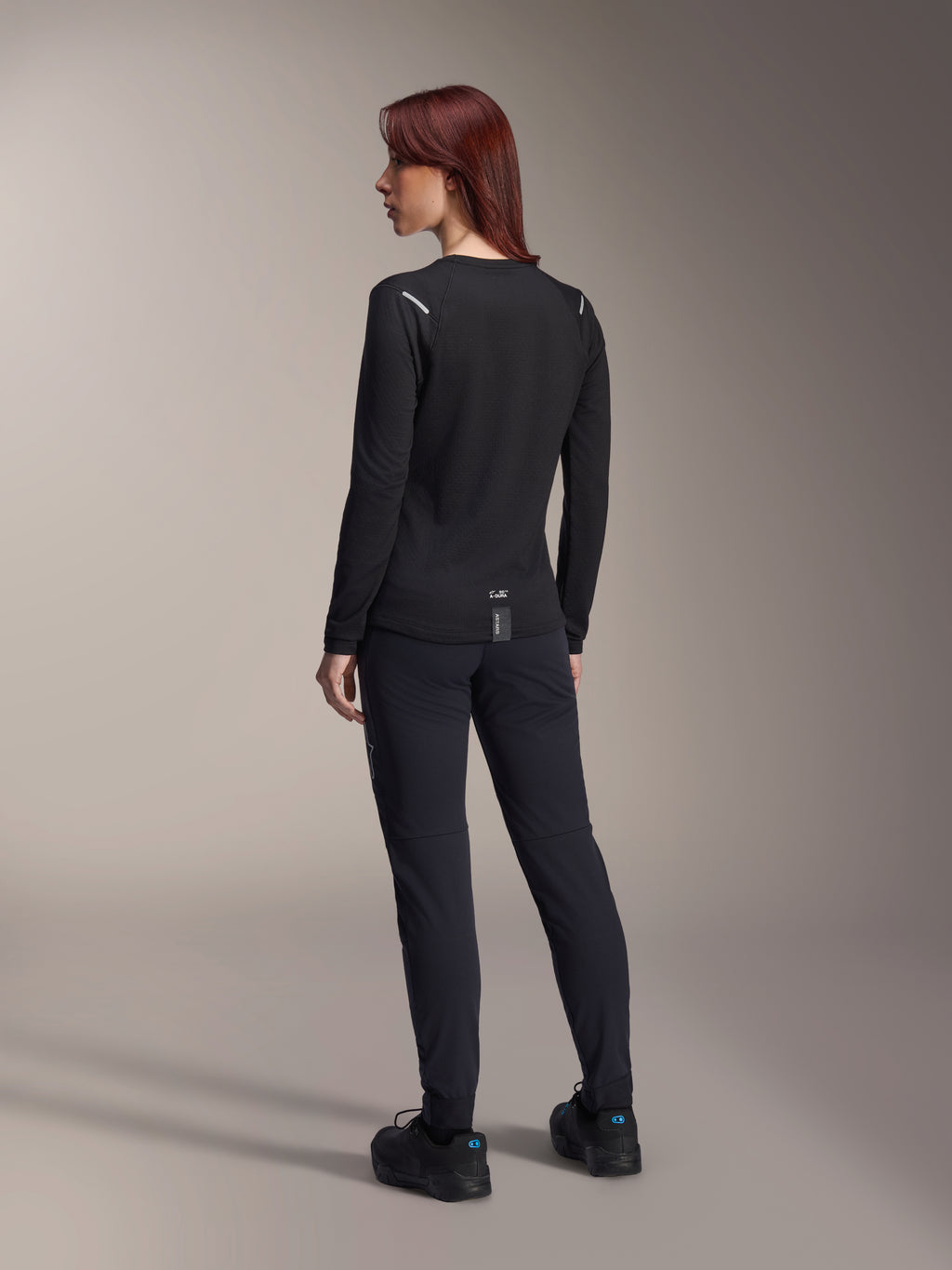 Alpinestars Stella A-Dura Thermal-tröja för damer, långärmad MTB-tröja i svart, vy bakifrån som visar reflekterande detaljer på axlarna och förlängt bakstycke, bärs med svarta byxor och skor, funktionskläder för mountainbike.