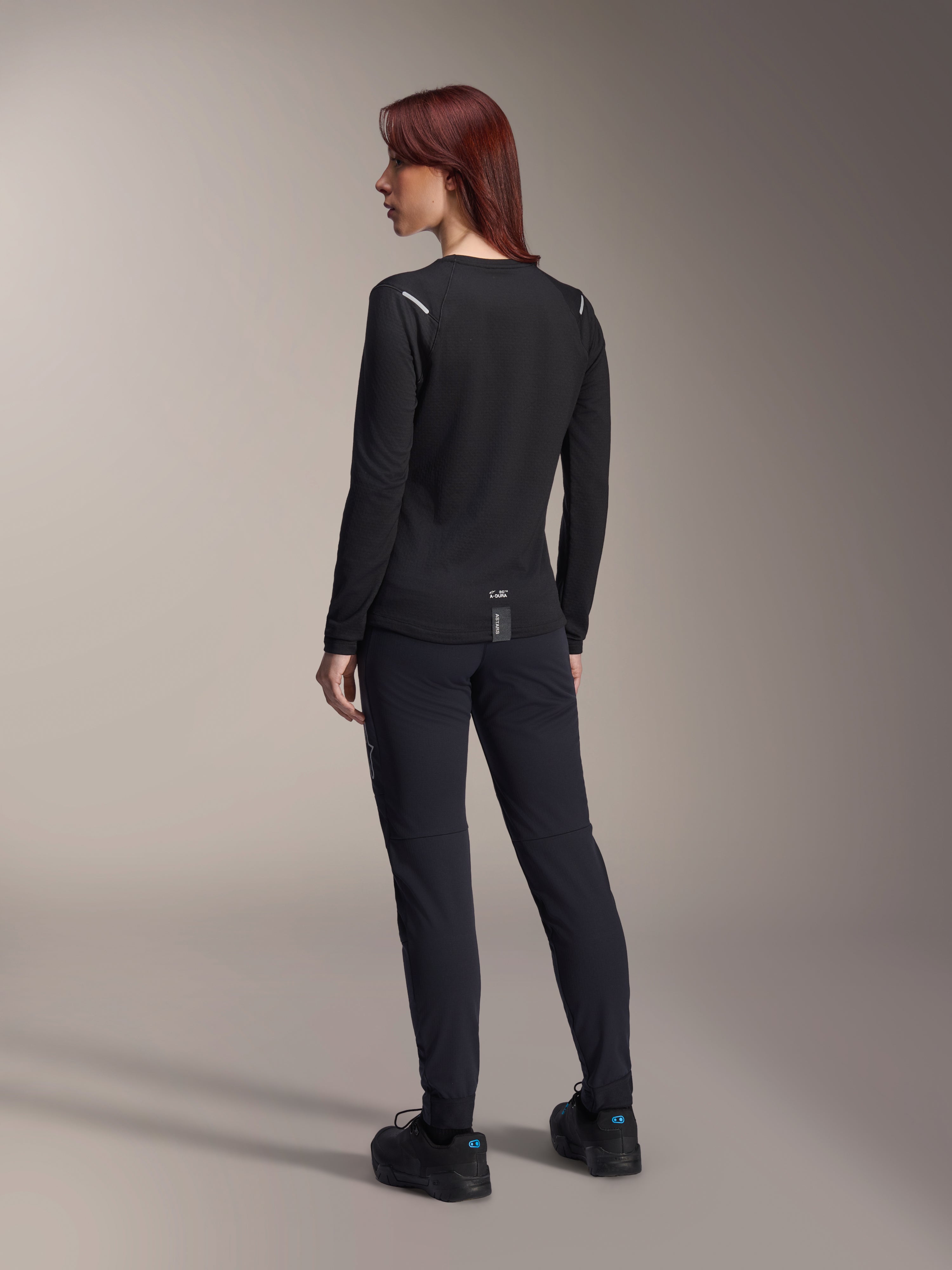 Alpinestars Stella A-Dura Thermal-tröja för damer, långärmad MTB-tröja i svart, vy bakifrån som visar reflekterande detaljer på axlarna och förlängt bakstycke, bärs med svarta byxor och skor, funktionskläder för mountainbike.