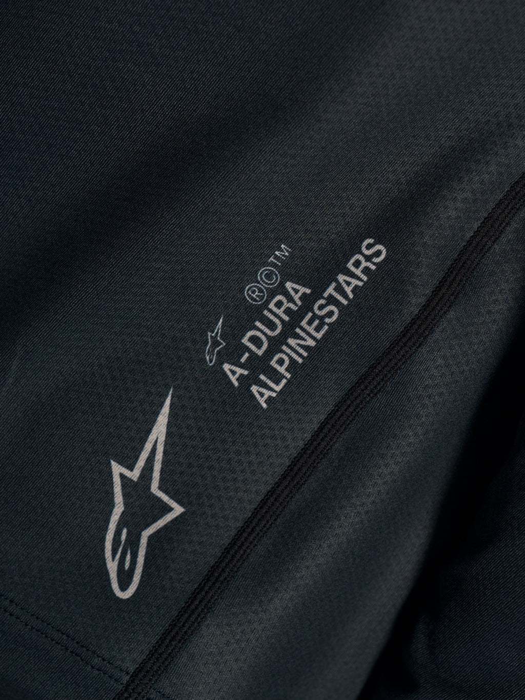 Svart Stella A-Dura Ride-tröja, närbild på tekniskt jacquardtyg med A-DURA ALPINESTARS och Astars-logotyper.