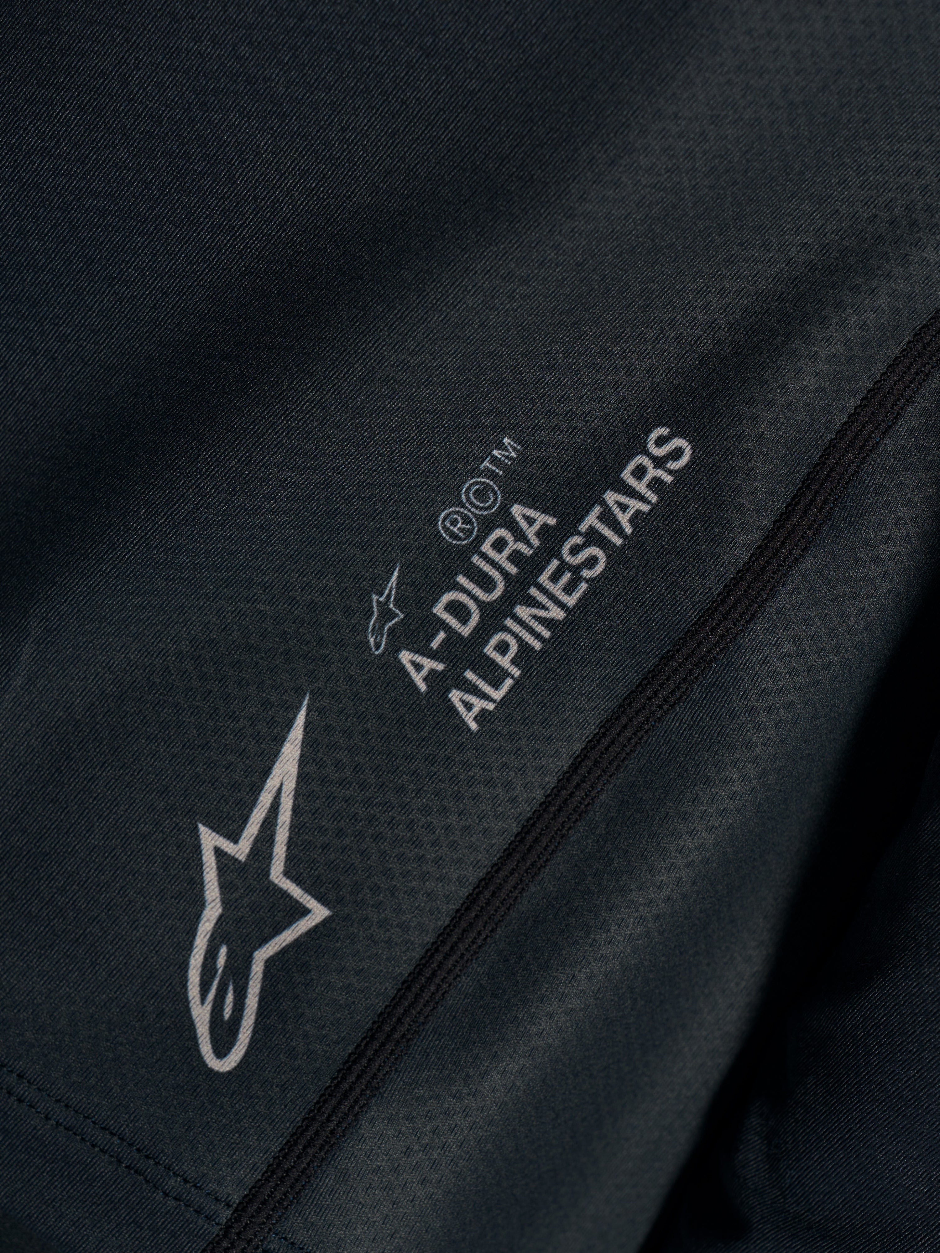 Svart Stella A-Dura Ride-tröja, närbild på tekniskt jacquardtyg med A-DURA ALPINESTARS och Astars-logotyper.