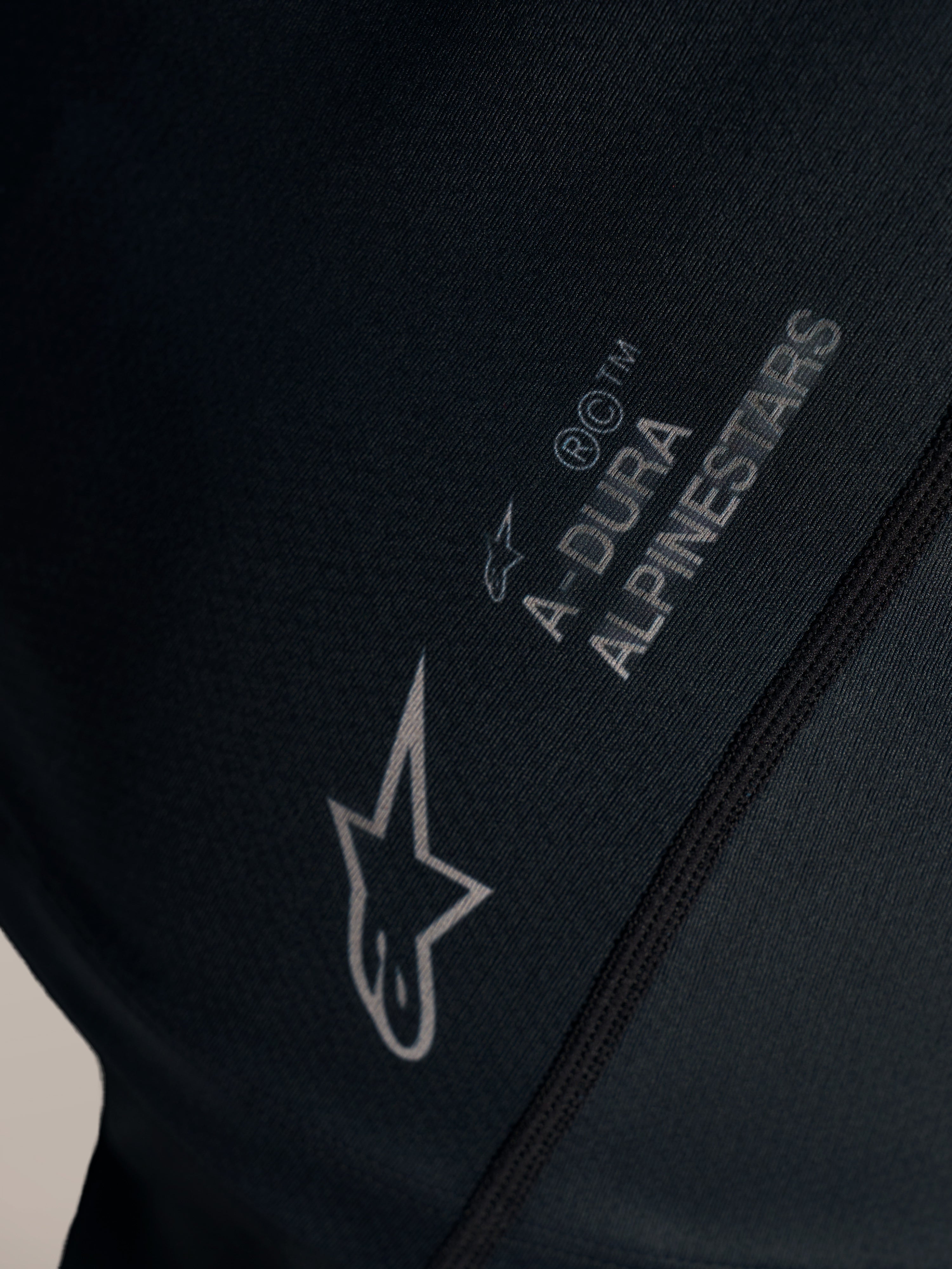 Närbild på svart Stella A-Dura Ride-tröja med vit Alpinestars-logotyp och A-DURA-märkning på jacquardvävt tyg.