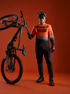 A-Dura ASTARS Långärmad Tröja 2.0 i orange-svart gradient med logotyp, visas med mountainbike-utrustning.