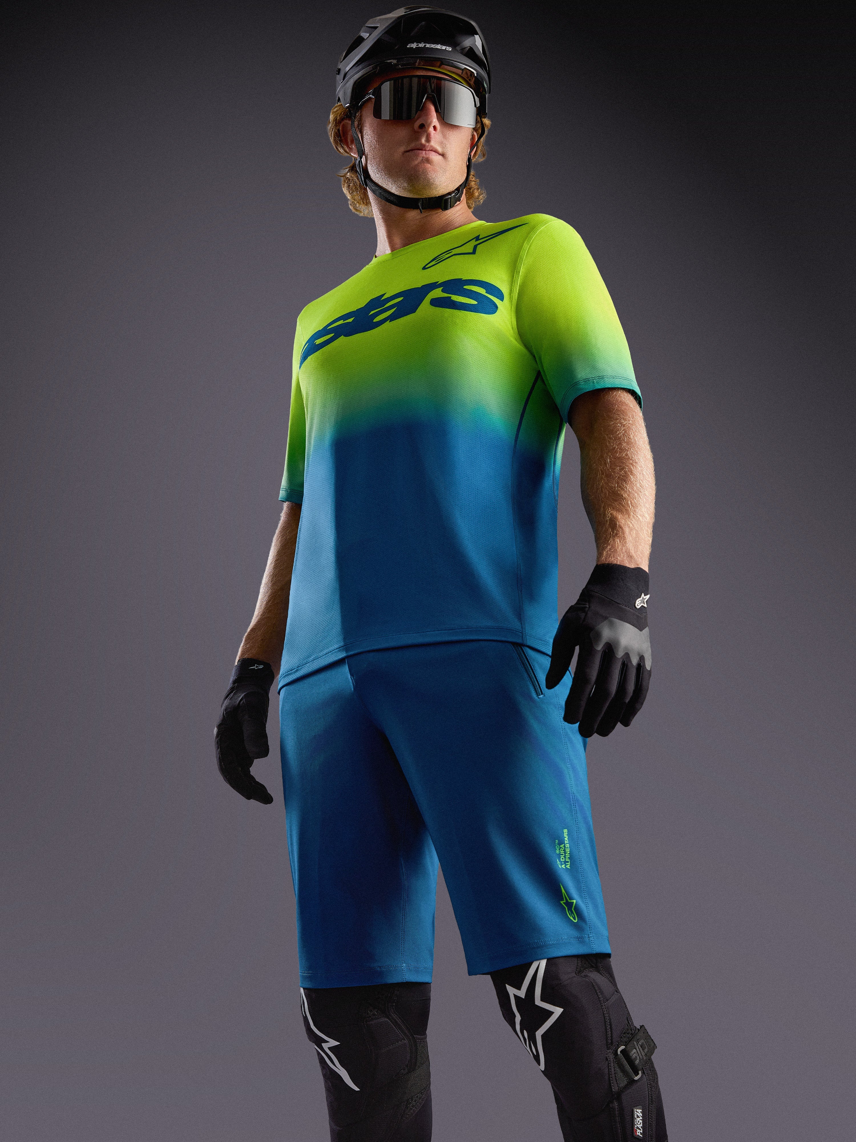 Alpinestars A-Dura ASTARS 2.0 MTB-tröja i blå och neongul gradient, matchande shorts och svart skyddsutrustning.