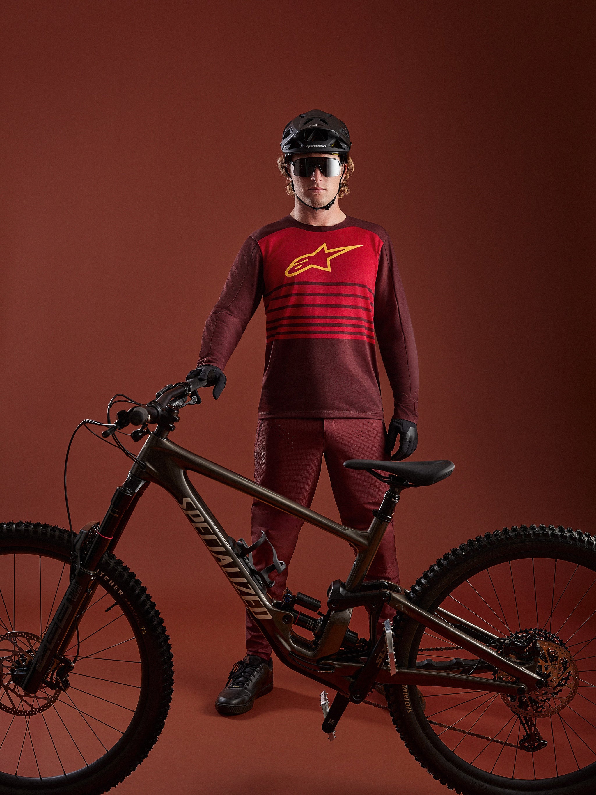 Alpinestars A-Dura Elite 2.0 långärmad MTB-tröja i bordeaux med röd bröstpanel och gul logotyp vid en mountainbike.