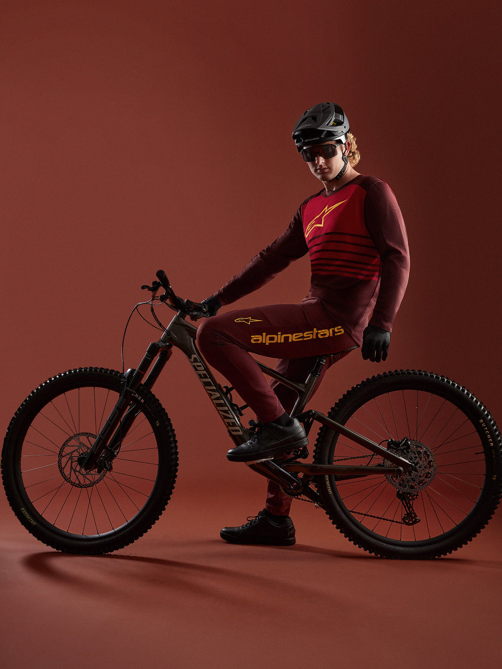 Alpinestars A-Dura Elite-set i bordeaux, tröja och byxor på en cyklist med Specialized-cykel i studiomiljö.