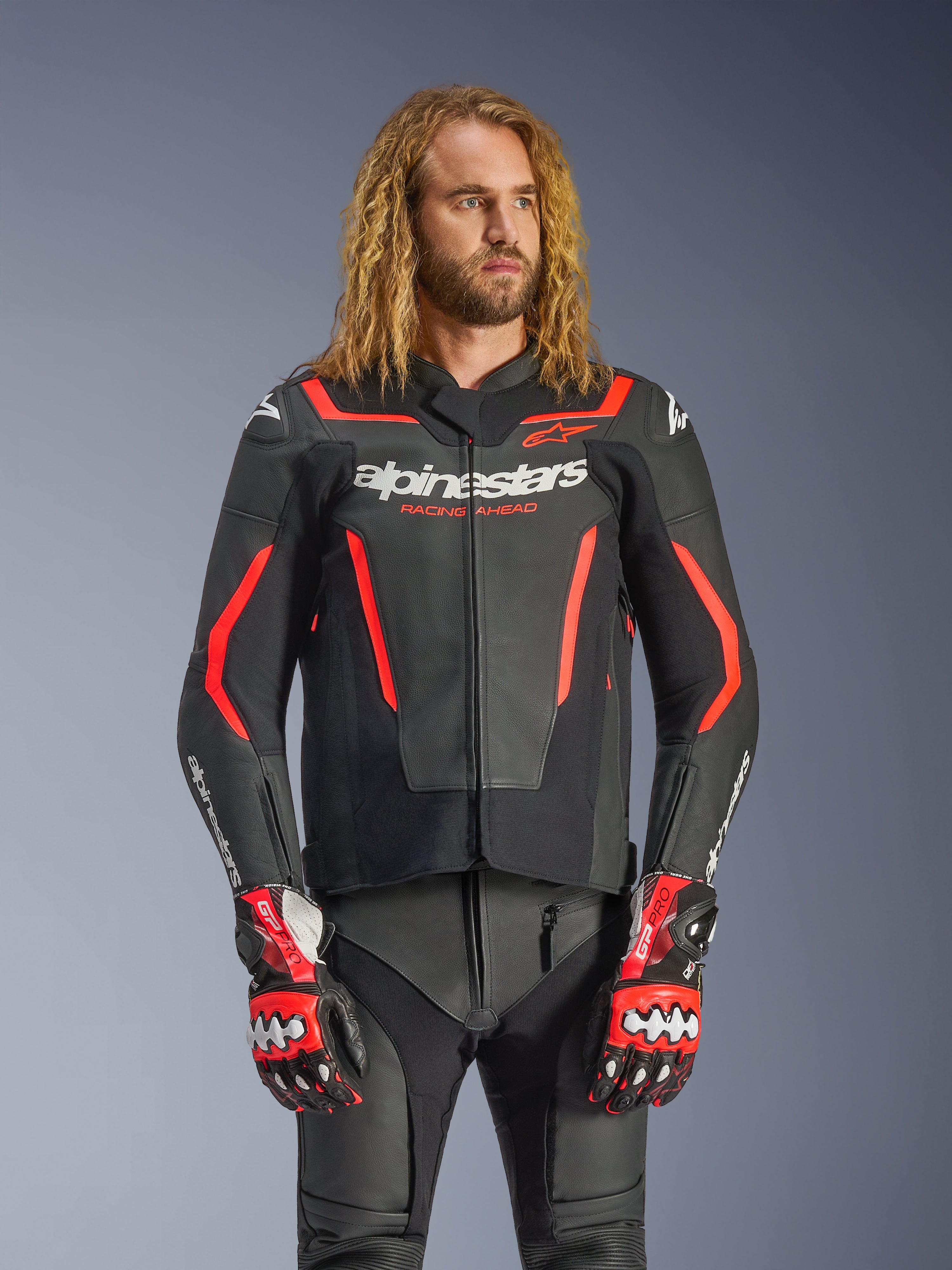 Alpinestars GP Force V2 läderjacka, sportmotorcykeljacka, svart och röd fluo, bärs av en manlig modell med långt vågigt hår, med vit Alpinestars-logotyp och texten "RACING AHEAD", kombinerad med matchande läderbyxor och handskar, professionell prestandautrustning