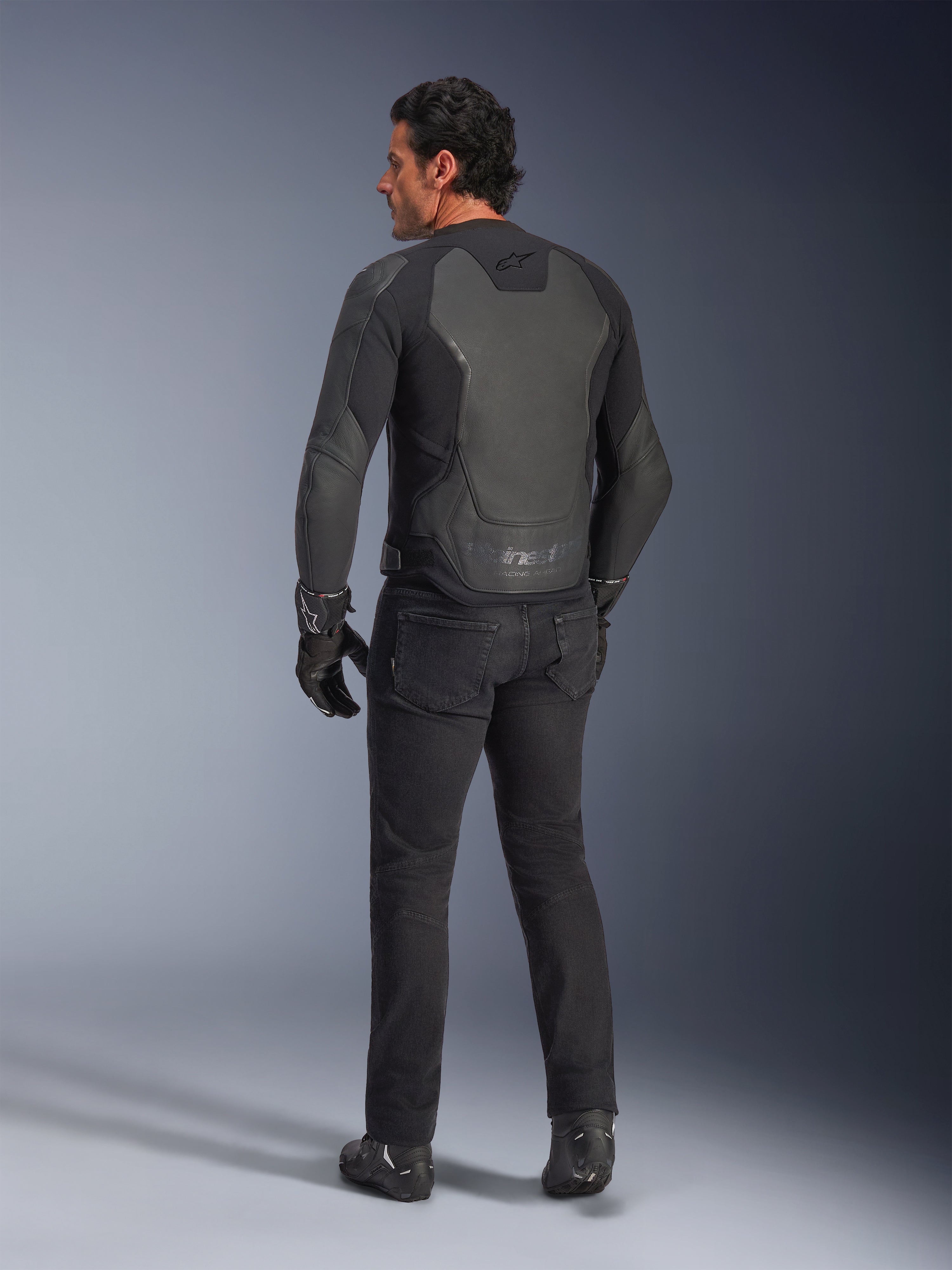 Rear view of a model wearing the Alpinestars GP Force V2 Leather Jacket in Black Black, featuring a multi-panel leather construction and sport fit, paired with black riding pants, gloves, and boots for a full motorcycle racing-inspired look.