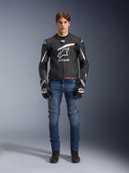 Male model wearing Alpinestars GP Plus R V4 Rideknit Leather Jacket in Black and White, styled with blue riding jeans, black motorcycle gloves, and black riding shoes, featuring a large white Astars logo on the chest, perforated leather panels, and white racing stripes on the sleeves, full-body standing view