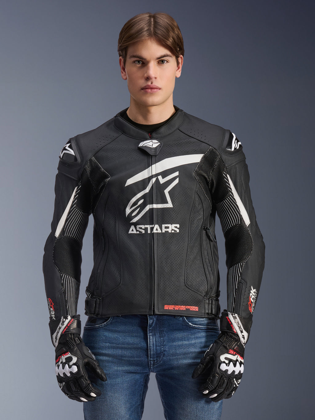 Alpinestars GP Plus R V4 Rideknit Leather Jacket, Black and White, motorcycle racing jacket worn by a male model with matching gloves and denim jeans, featuring a prominent white ASTARS logo on the chest, perforated leather panels, and protective shoulder sliders