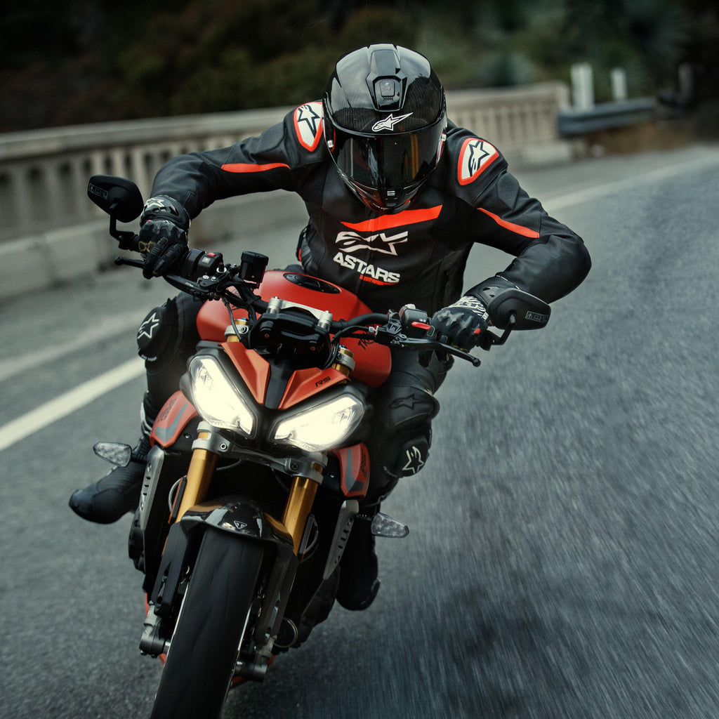 Motorcyklist som kör en röd sportmotorcykel, Alpinestars GP Plus R V4 Rideknit läderjacka, svart med fluorescerande röda och vita detaljer, med vit Alpinestars-logotyp och 'ASTARS'-text, matchat med svart racinghjälm och handskar, högpresterande körutrustning i aktion på en väg.