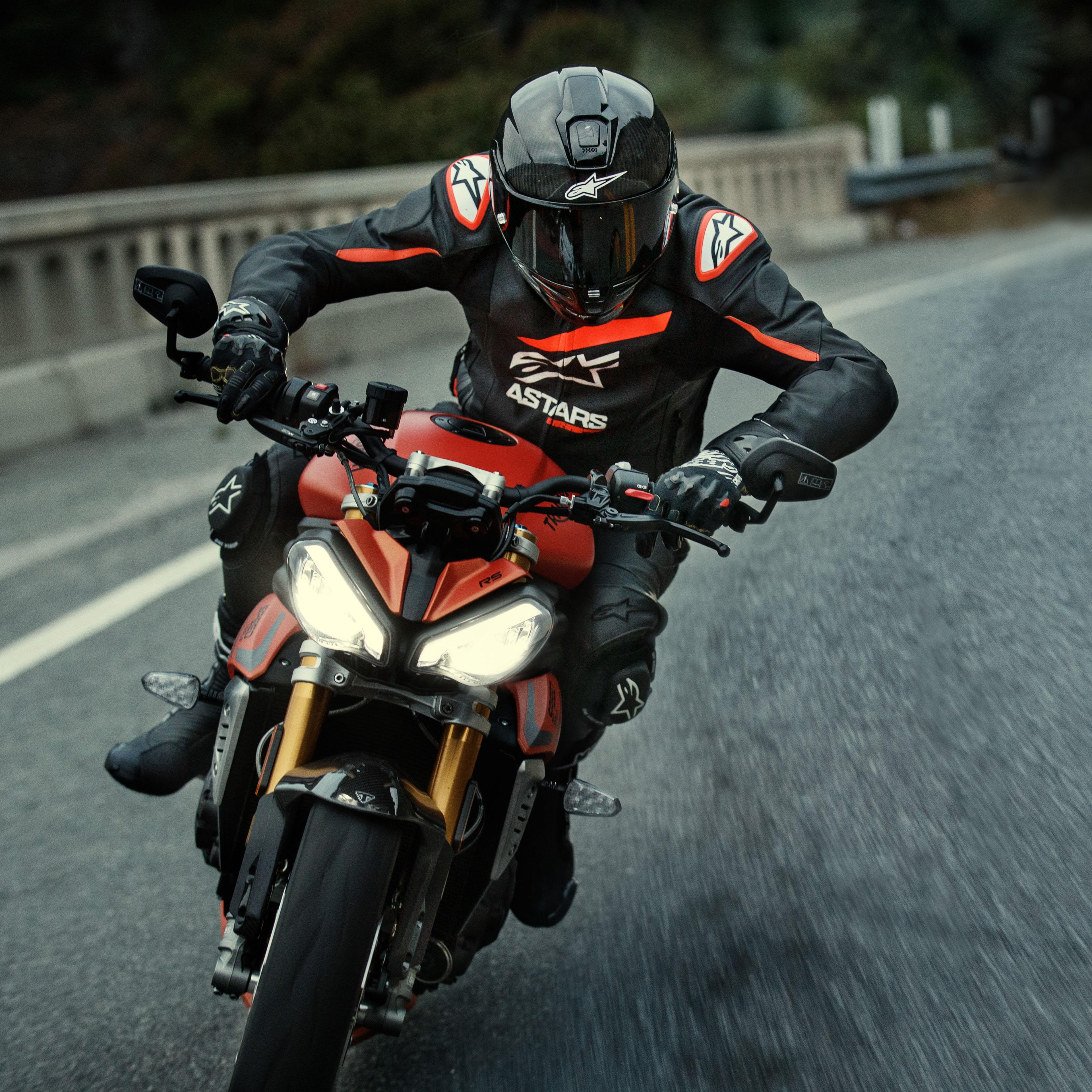 Motorcyklist som kör en röd sportmotorcykel, Alpinestars GP Plus R V4 Rideknit läderjacka, svart med fluorescerande röda och vita detaljer, med vit Alpinestars-logotyp och 'ASTARS'-text, matchat med svart racinghjälm och handskar, högpresterande körutrustning i aktion på en väg.
