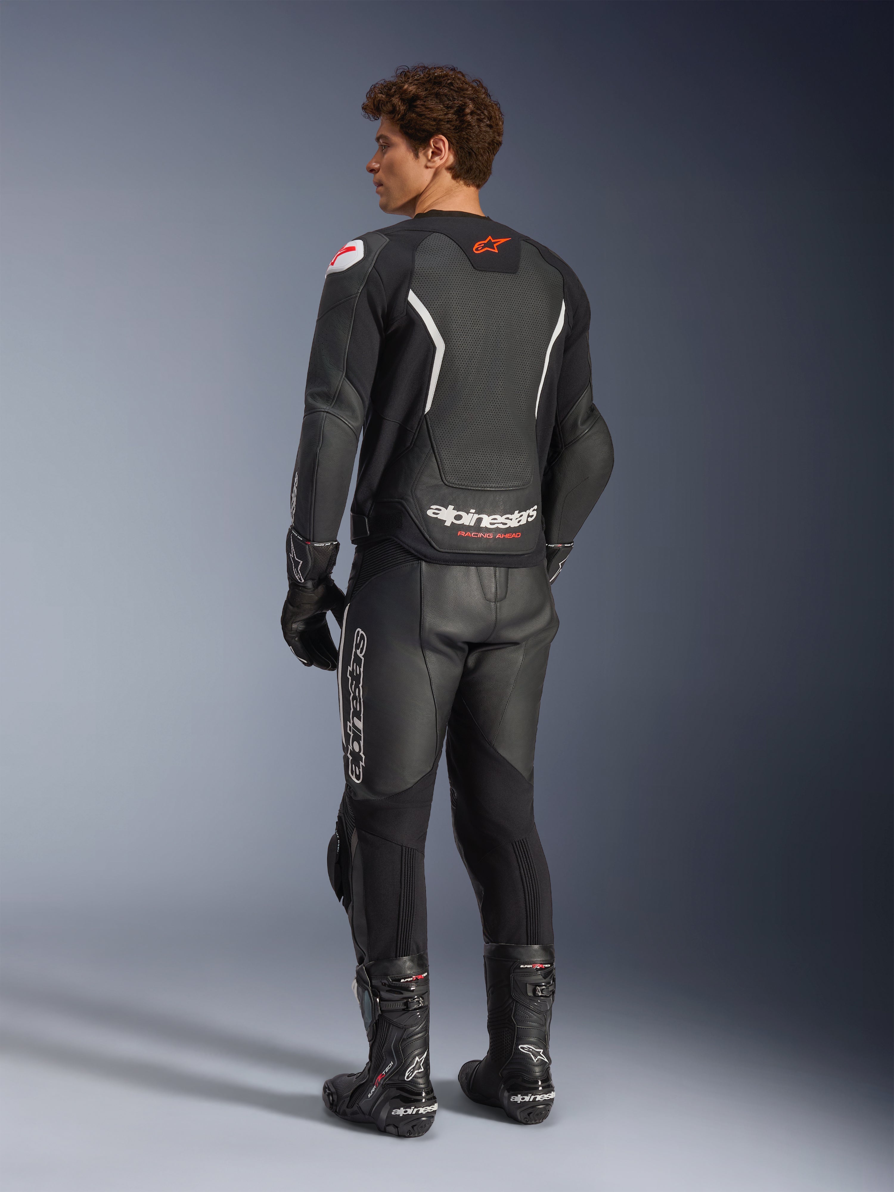 Alpinestars GP Force V2 Air läderjacka, svart och vit, vy bakifrån på modell med en perforerad ryggpanel för ventilation, vit logotyp och röd stjärnlogotyp, buren med matchande racingbyxor i läder, handskar och stövlar
