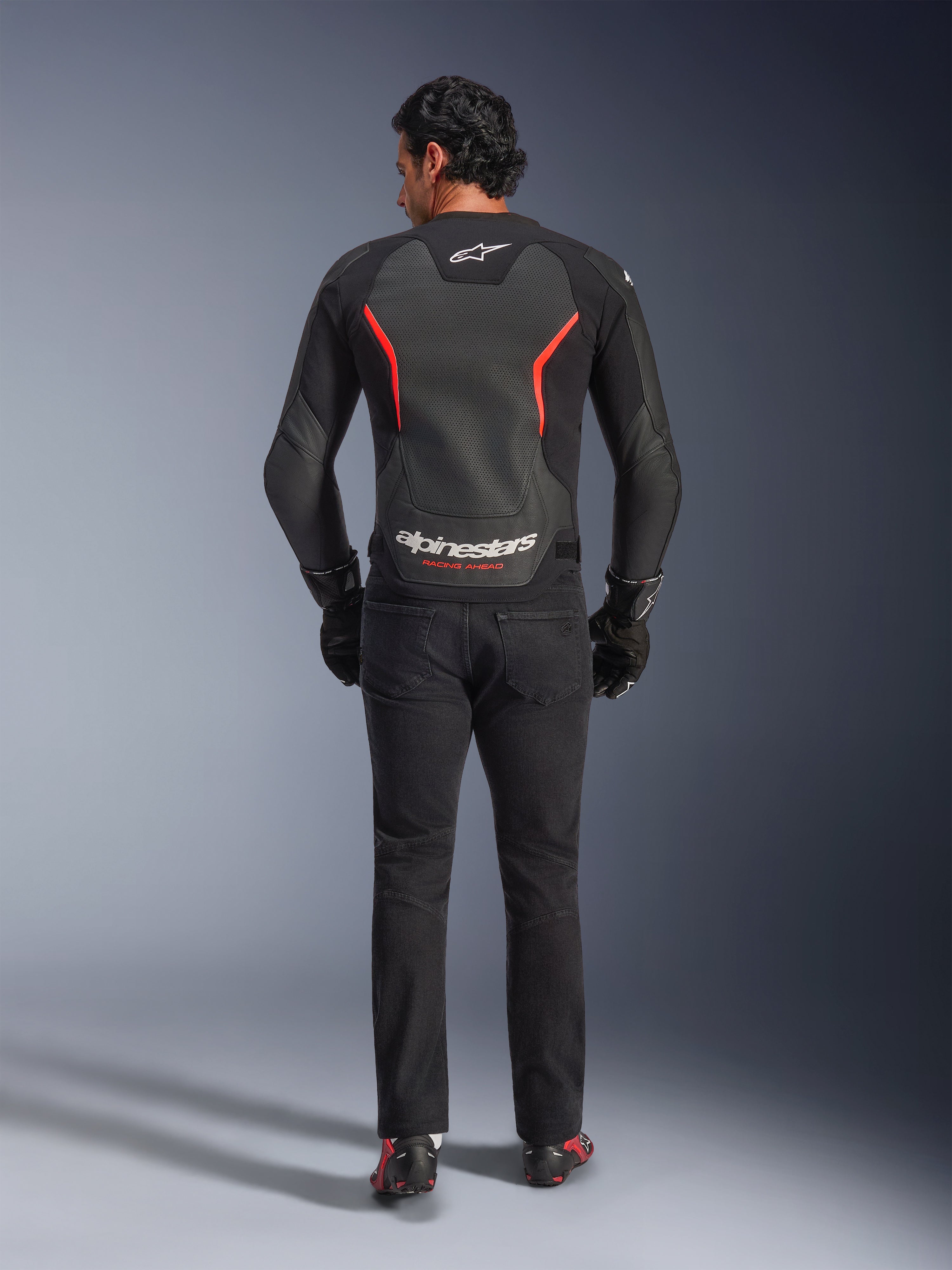 Alpinestars GP Force V2 Air Leather Jacket in Black and Red Fluo, rear view on model, featuring perforated leather back panels, red accents, and Alpinestars branding, styled with black riding jeans and gloves for a sport motorcycle look.