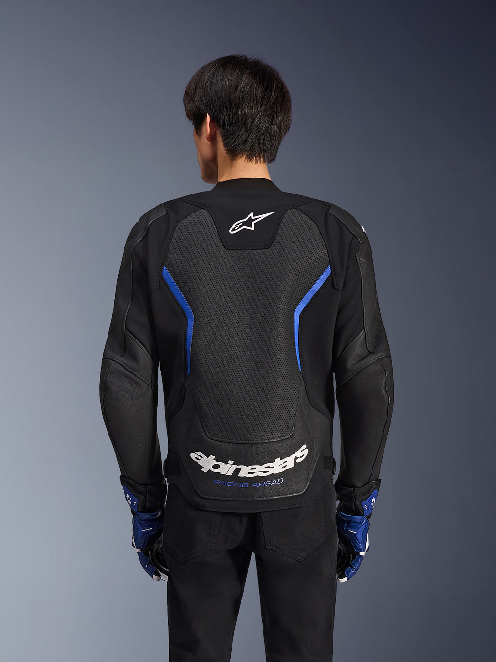 Alpinestars GP Force V2 Airflow läderjacka, Black Metallic Blue, vy bakifrån, med en stor perforerad ryggpanel för ventilation, blå detaljer och vit Alpinestars-logotyp, bärs med matchande blå handskar och svarta byxor för sportkörning.
