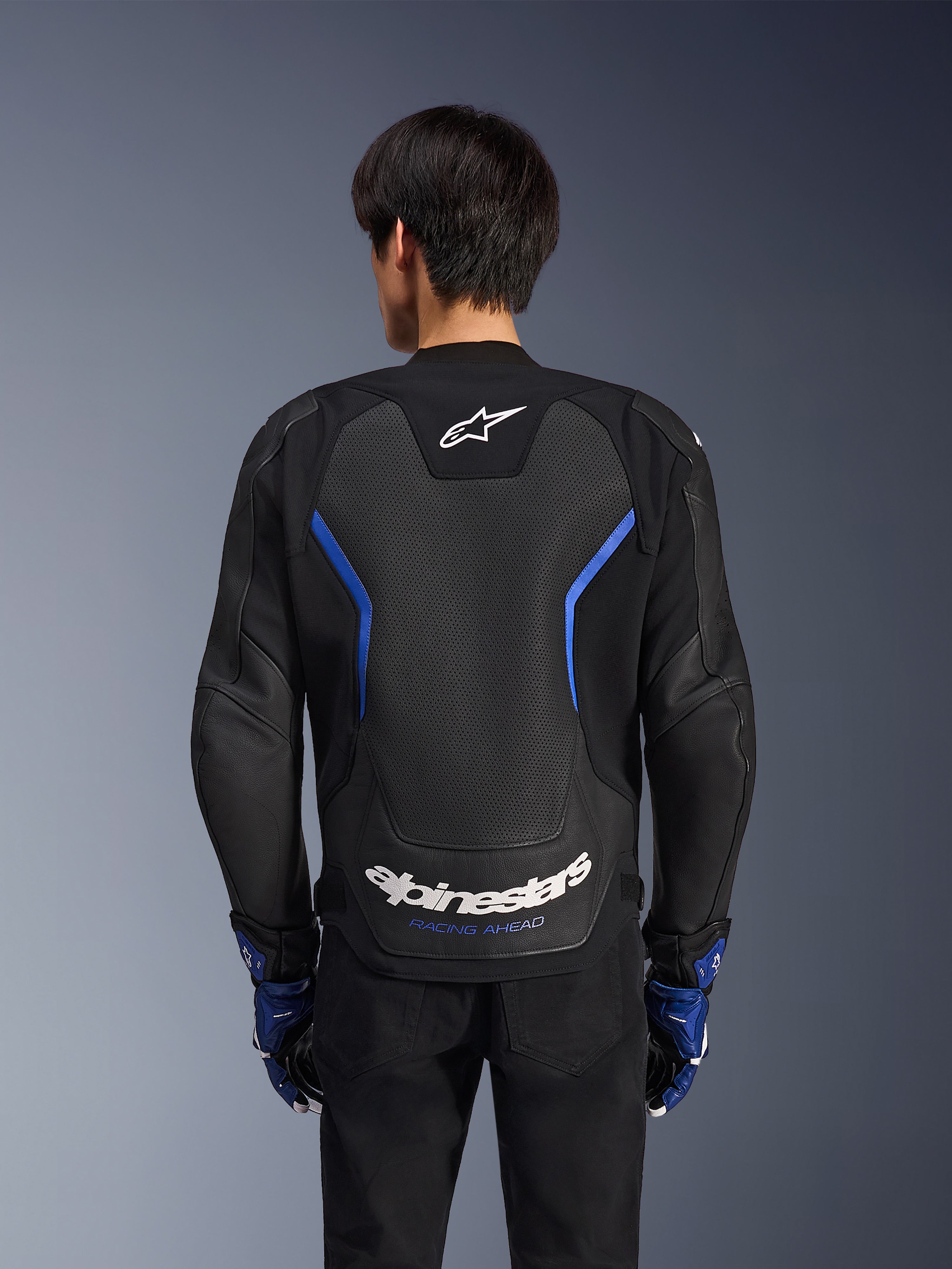 Alpinestars GP Force V2 Airflow läderjacka, Black Metallic Blue, vy bakifrån, med en stor perforerad ryggpanel för ventilation, blå detaljer och vit Alpinestars-logotyp, bärs med matchande blå handskar och svarta byxor för sportkörning.