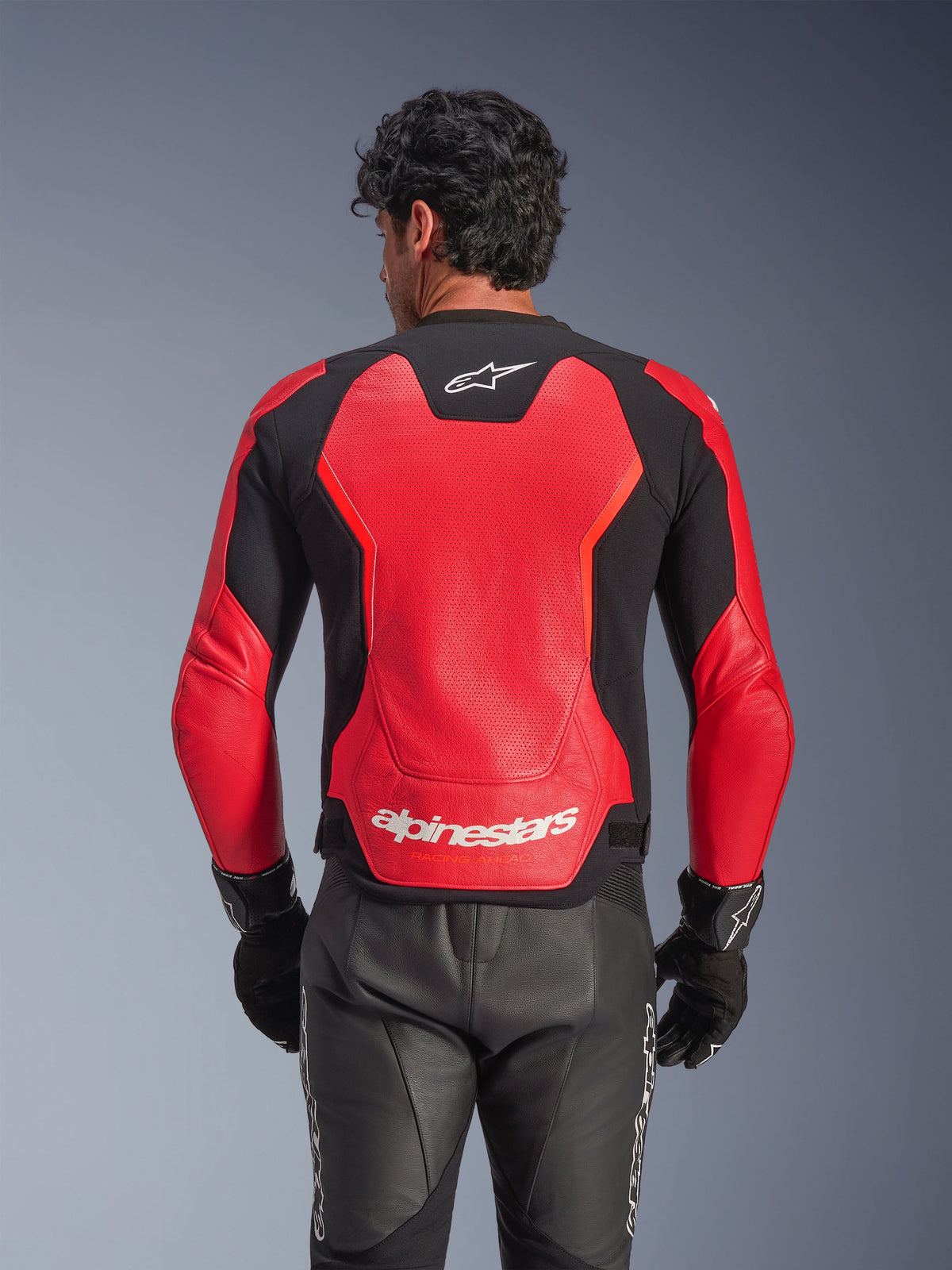 Alpinestars GP Force V2 Air läderjacka i rött och svart, vy bakifrån som visar det perforerade läderchassit för luftflöde, vit Alpinestars-logotyp på nedre delen av ryggen och integrerade stretchpaneler, bärs med svarta motorcykelbyxor i läder och handskar.