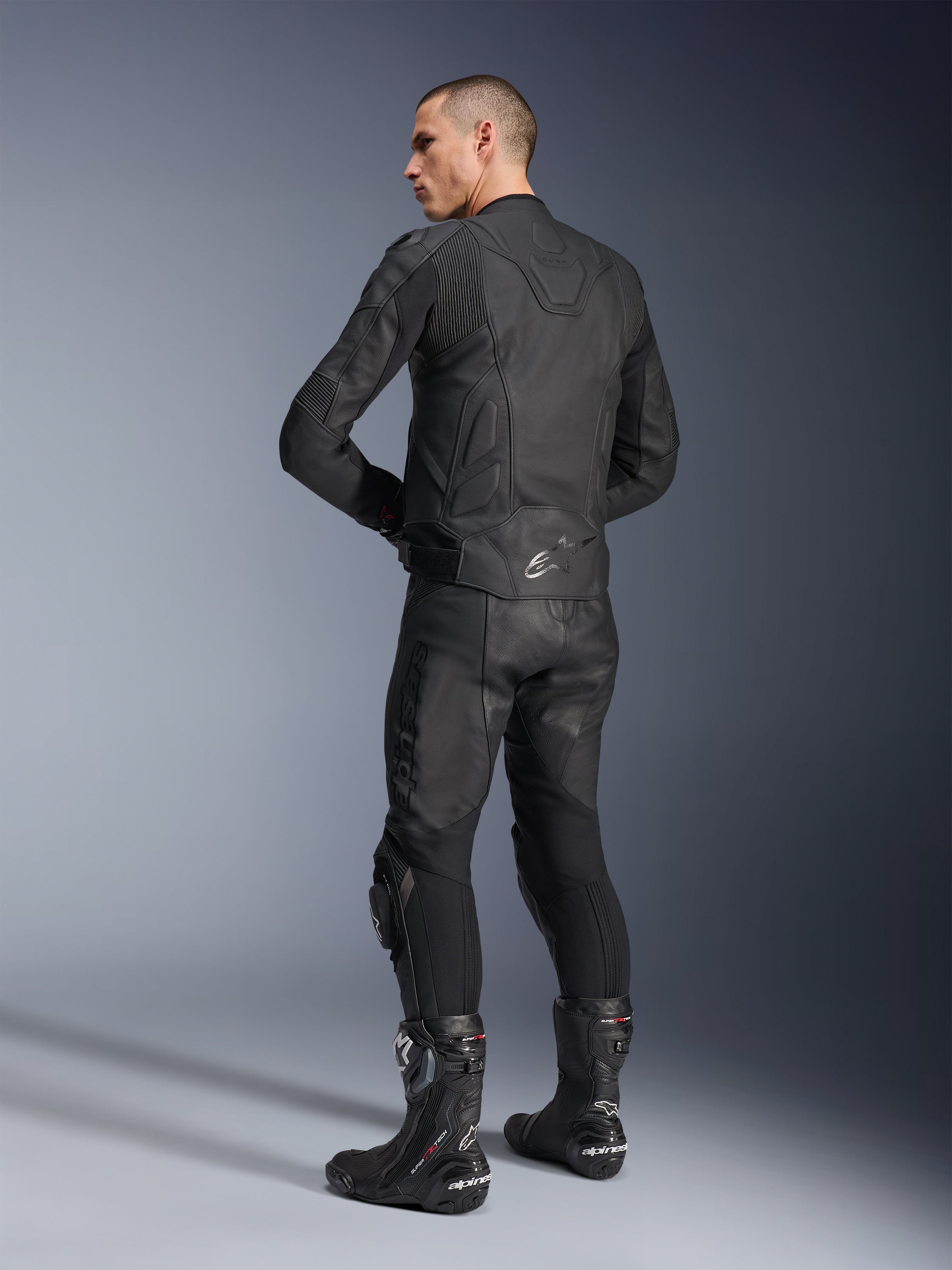 Alpinestars Dusk Leather Jacket, Black, Rear three-quarter view on a model wearing matching leather motorcycle pants and performance racing boots, featuring leather accordion stretch panels, silver Alpinestars star logo on the lower back, and DUSK branding on the upper back, professional studio shot