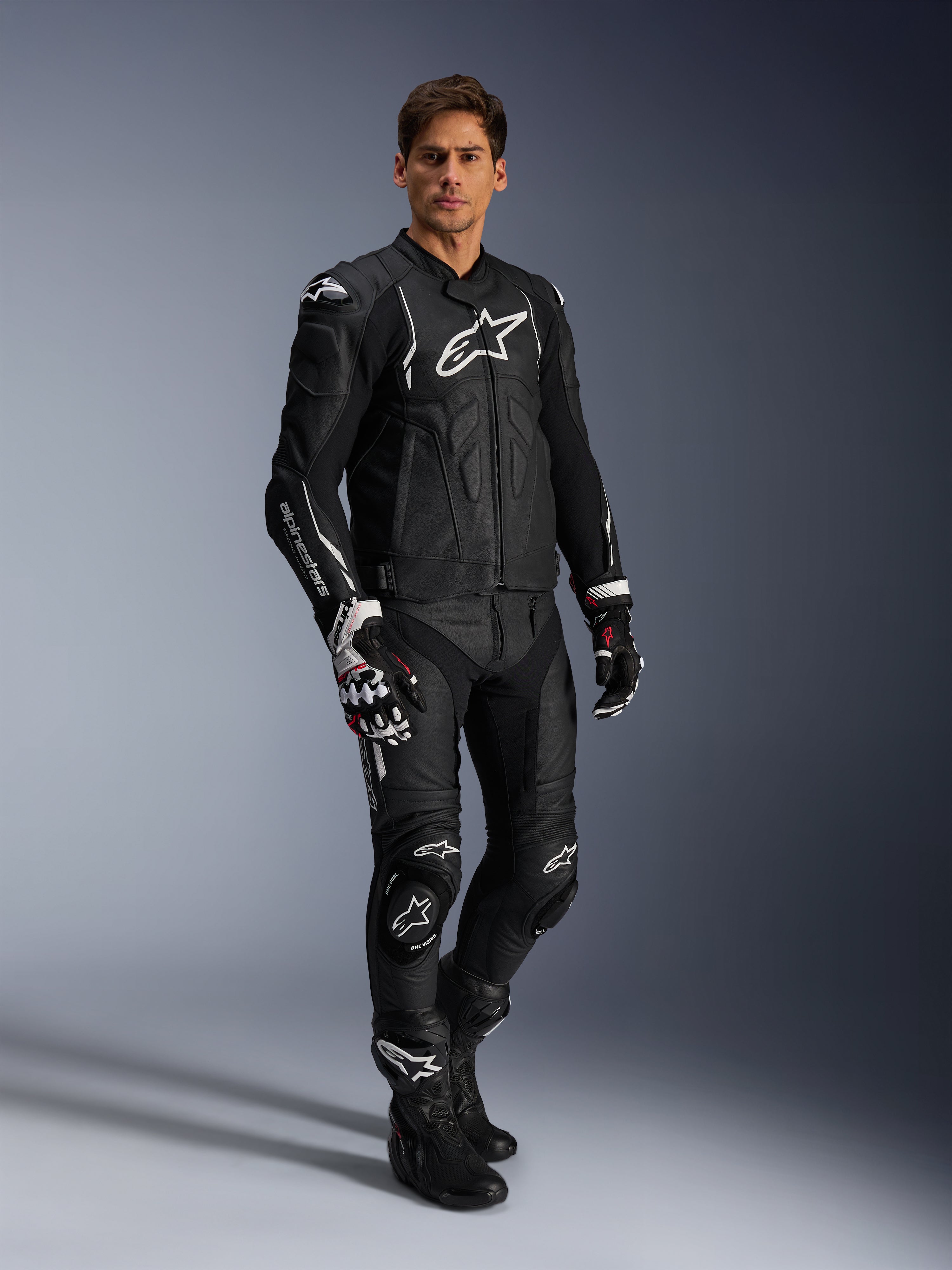 Male rider wearing the Alpinestars Dusk Leather Jacket in Black and White, premium leather jacket with large white branding on the chest, paired with matching leather racing pants, gloves, and boots, complete performance sport riding gear.