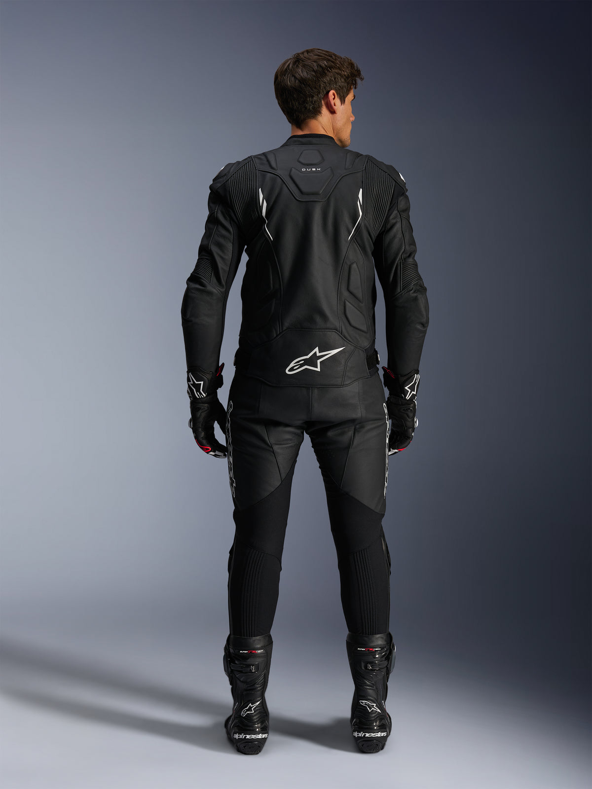 Motorcykelförare sedd bakifrån i Alpinestars Dusk läderjacka och Missile V3 läderbyxor, helsvart läderställ med vita detaljer, jackan har ”Dusk”-märkning och Alpinestars-logotyp på ryggen, matchas med svarta racingstövlar och handskar, koordinerad motorsportoutfit för roadracing.