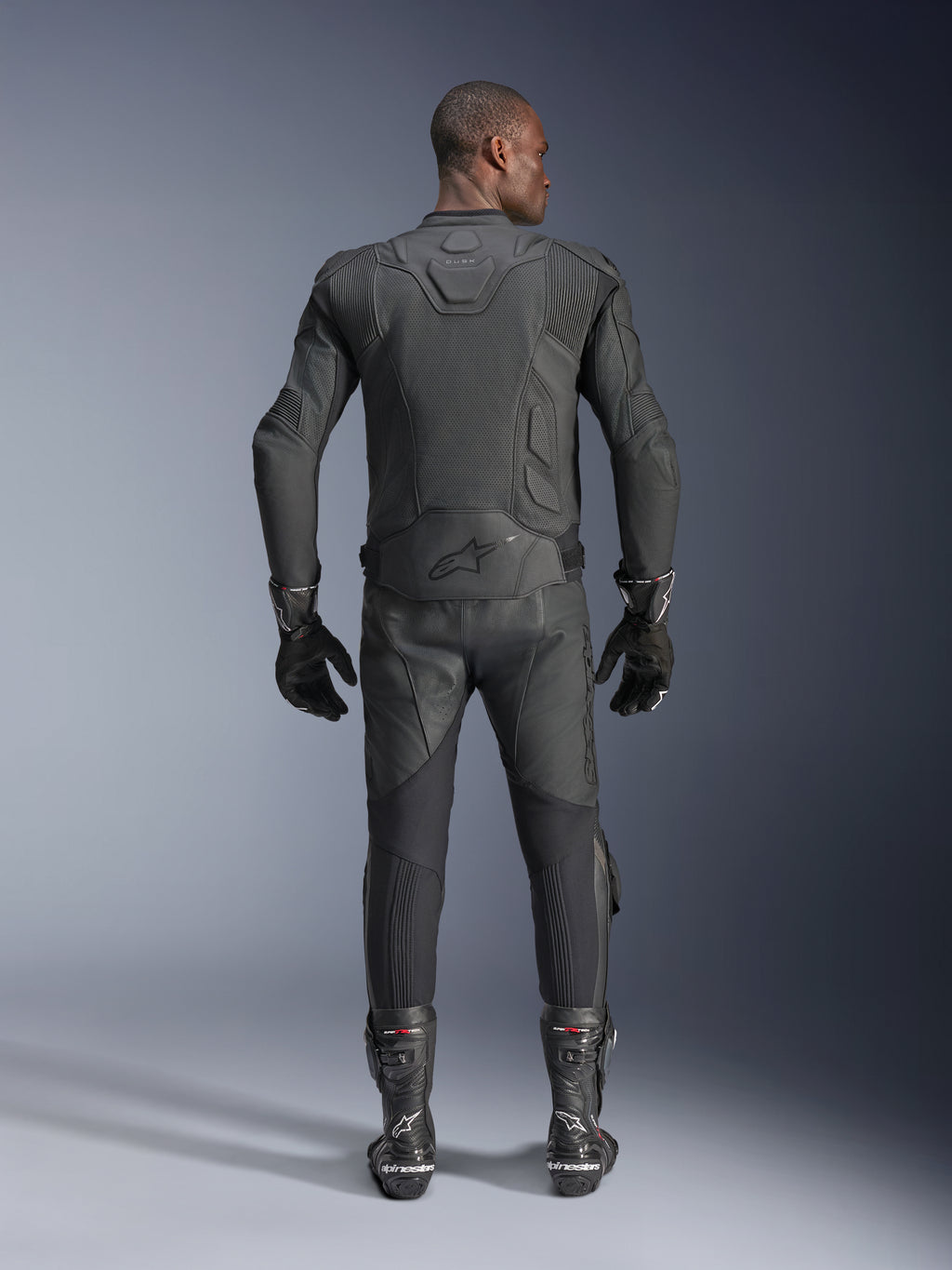 Manlig modell sedd bakifrån i Alpinestars Dusk Airflow läderjacka i svart, matchad med svarta racingbyxor i läder, handskar och stövlar, med omfattande perforeringar för ventilation och Alpinestars-logotypen på nedre delen av ryggen.