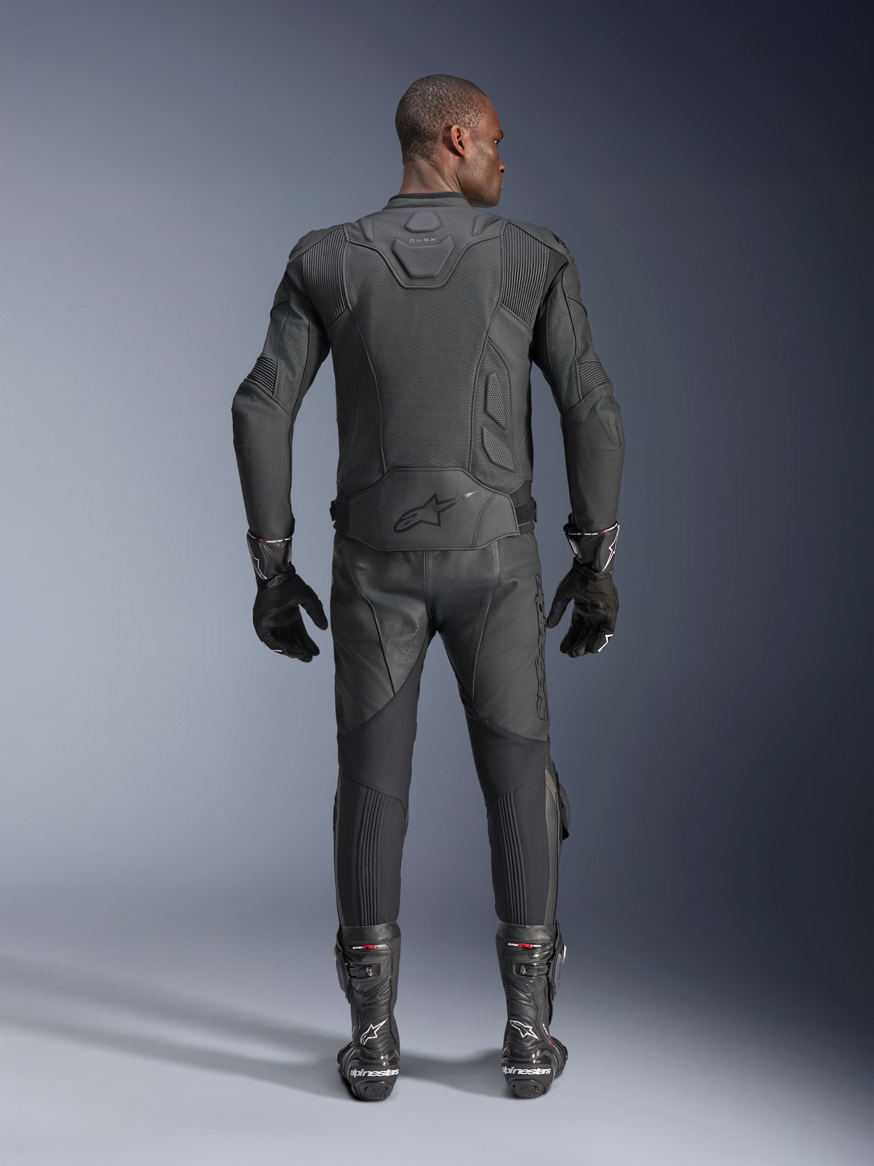 Manlig modell sedd bakifrån i Alpinestars Dusk Airflow läderjacka i svart, matchad med svarta racingbyxor i läder, handskar och stövlar, med omfattande perforeringar för ventilation och Alpinestars-logotypen på nedre delen av ryggen.