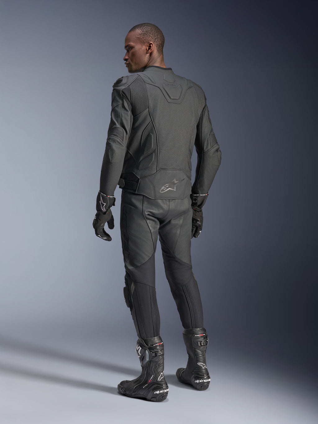 Manlig förare sedd bakifrån klädd i Alpinestars Dusk Airflow Läderjacka i Black Black, kombinerad med matchande läderbyxor, handskar och stövlar, vilket framhäver de omfattande ryggperforeringarna och den sportiga passformen.