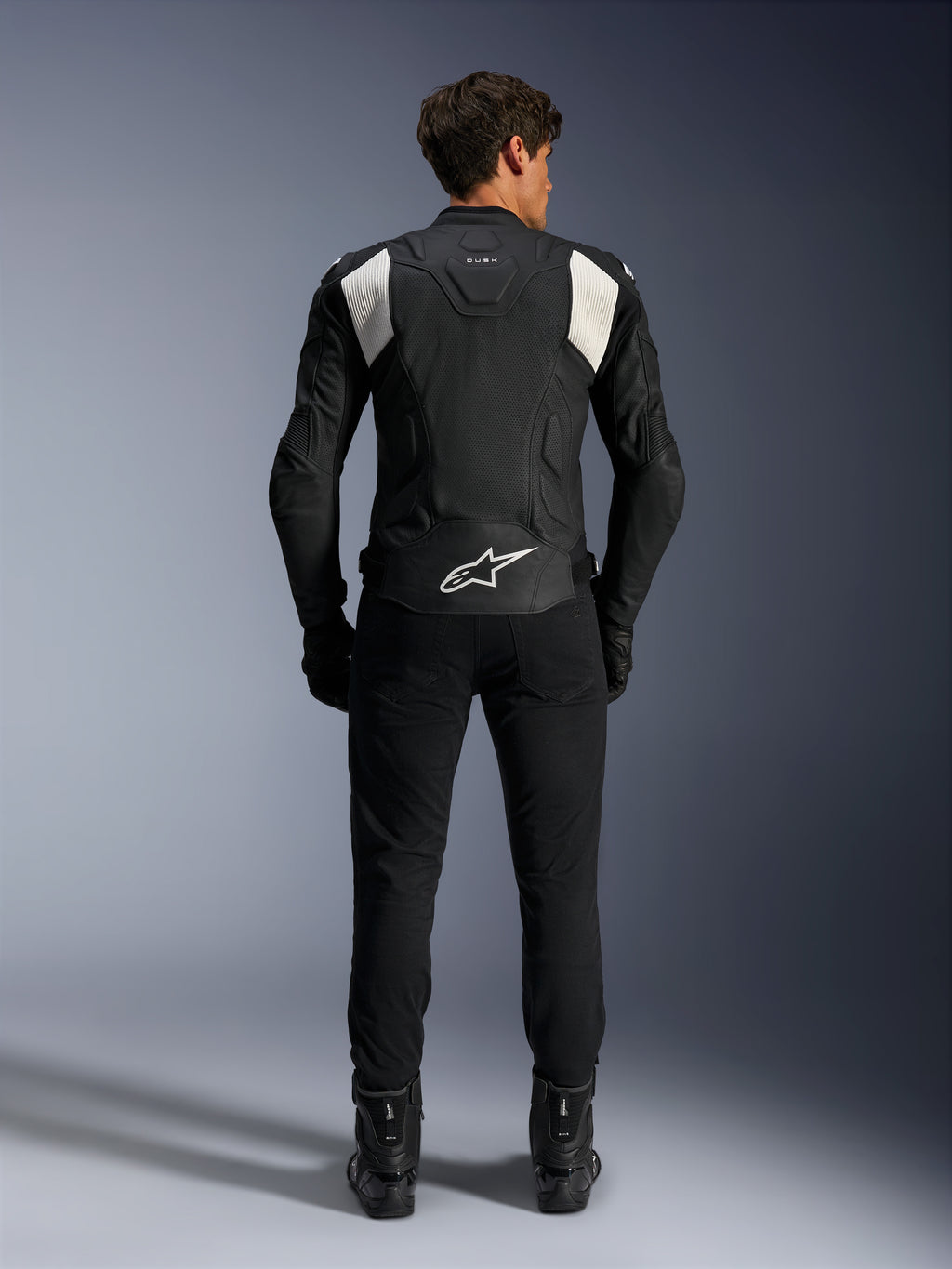 Alpinestars Dusk Airflow Läderjacka, svart och vit, vy bakifrån av en förare i svarta byxor och stövlar, med perforerat läder för ventilation, vita dragspelspaneler på axlarna och vit logotyp på nedre delen av ryggen