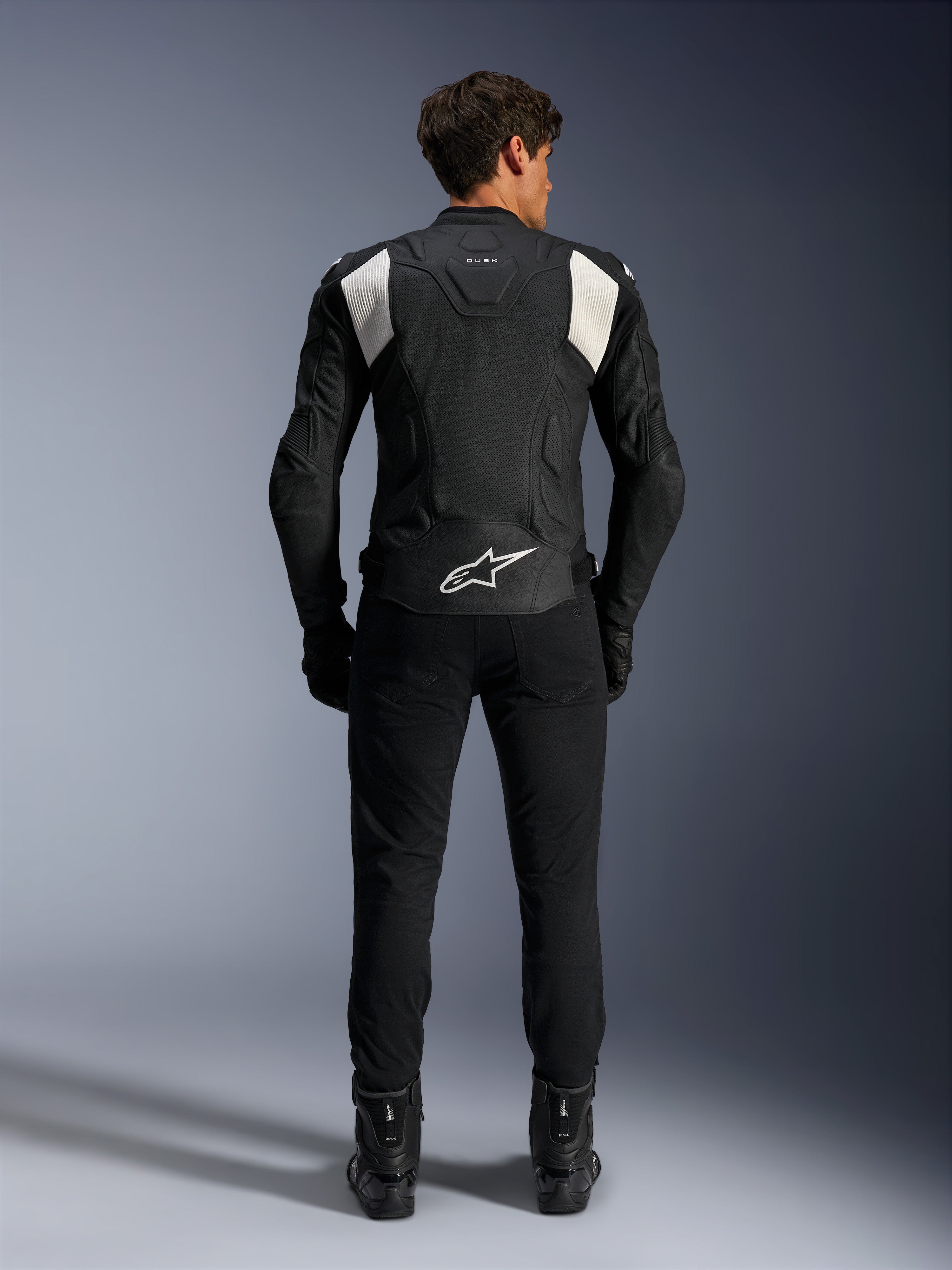 Alpinestars Dusk Airflow Läderjacka, svart och vit, vy bakifrån av en förare i svarta byxor och stövlar, med perforerat läder för ventilation, vita dragspelspaneler på axlarna och vit logotyp på nedre delen av ryggen