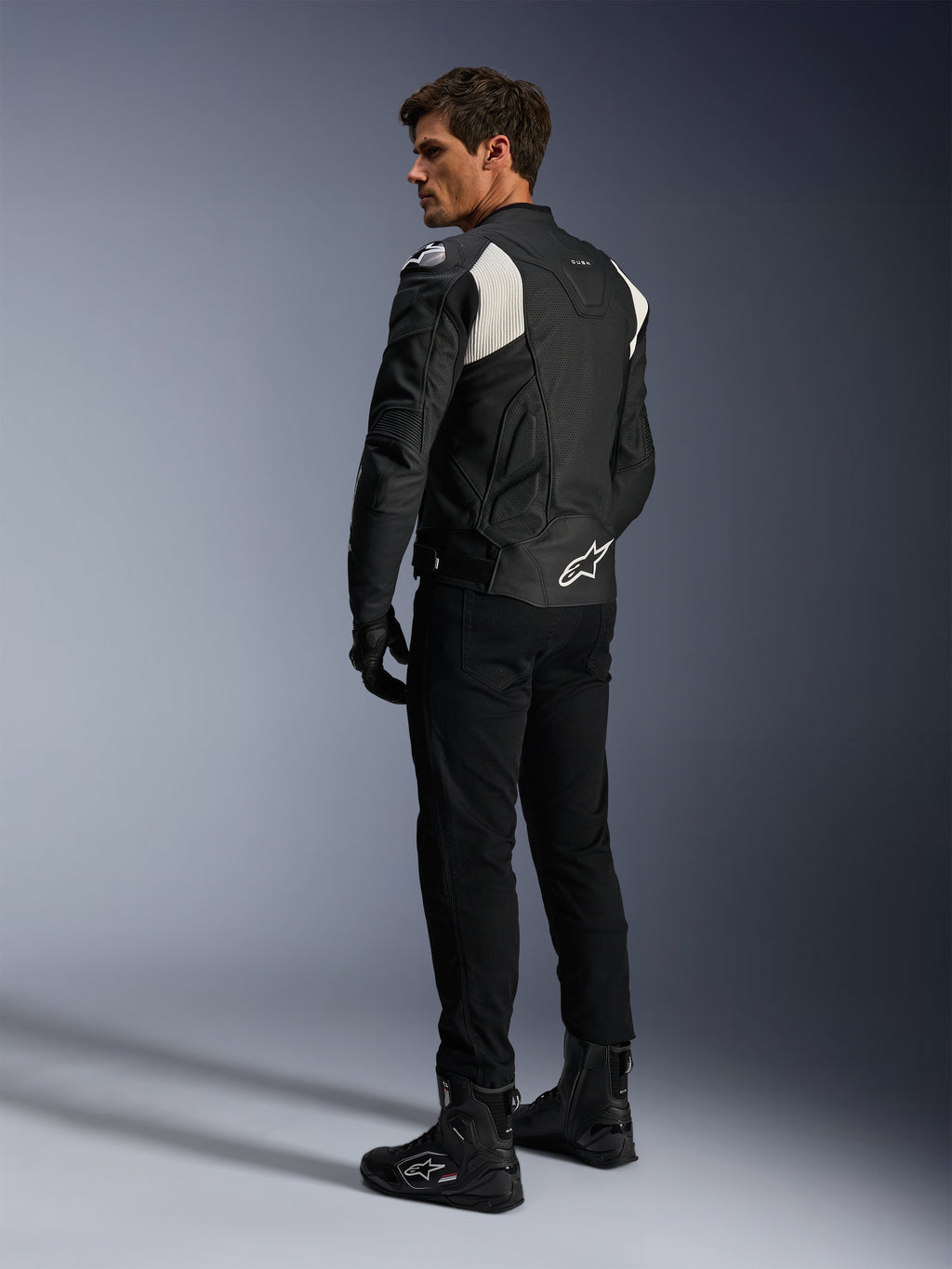 Alpinestars Dusk Airflow skinnjacka, svart och vit, vy bakifrån på modell som bär den perforerade skinnjackan med matchande svarta byxor och stövlar, vita dragspelsstretchpaneler och en tydlig logotyp på nedre delen av ryggen, sportig motorcykelutrustning
