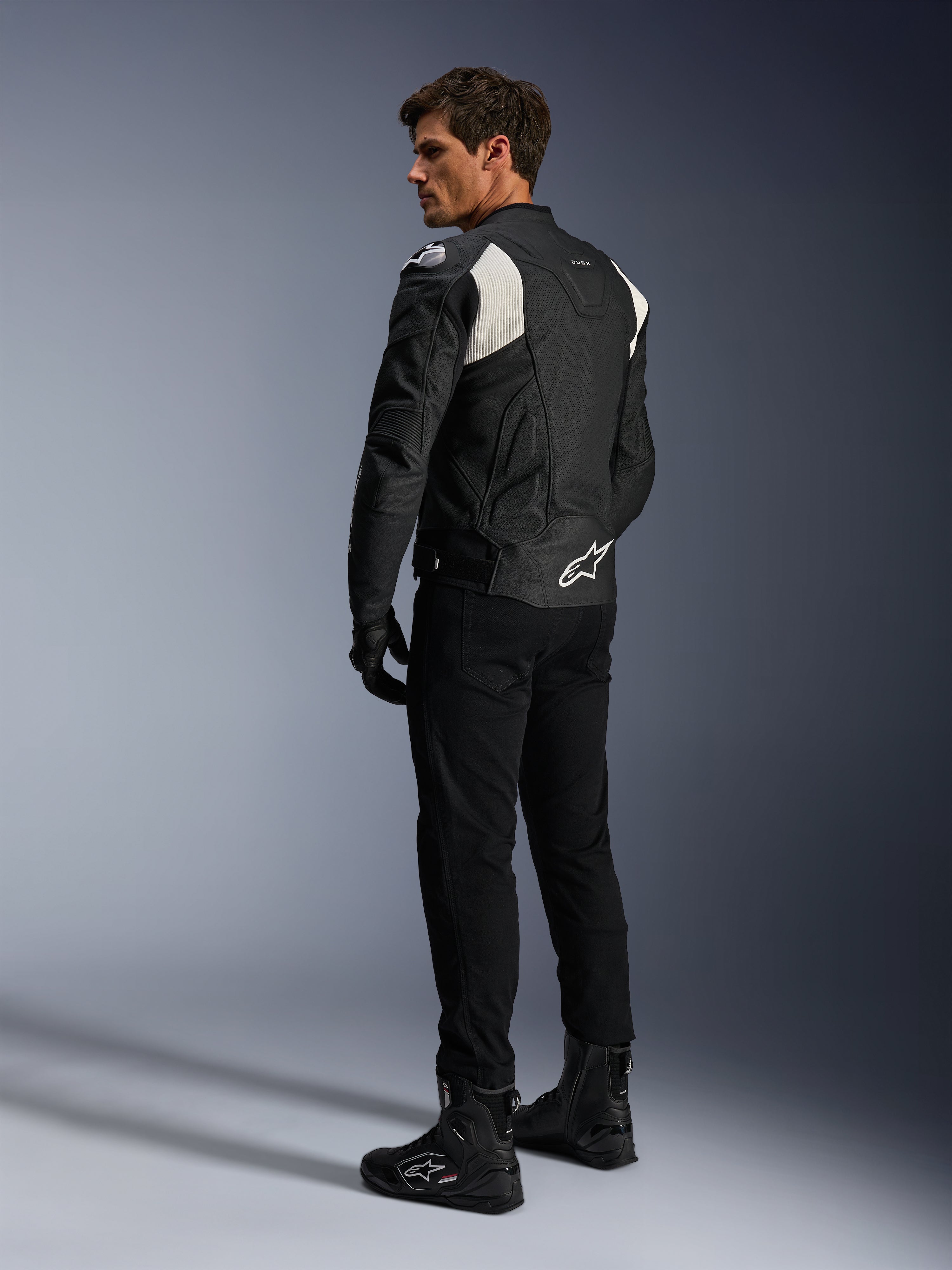 Alpinestars Dusk Airflow skinnjacka, svart och vit, vy bakifrån på modell som bär den perforerade skinnjackan med matchande svarta byxor och stövlar, vita dragspelsstretchpaneler och en tydlig logotyp på nedre delen av ryggen, sportig motorcykelutrustning