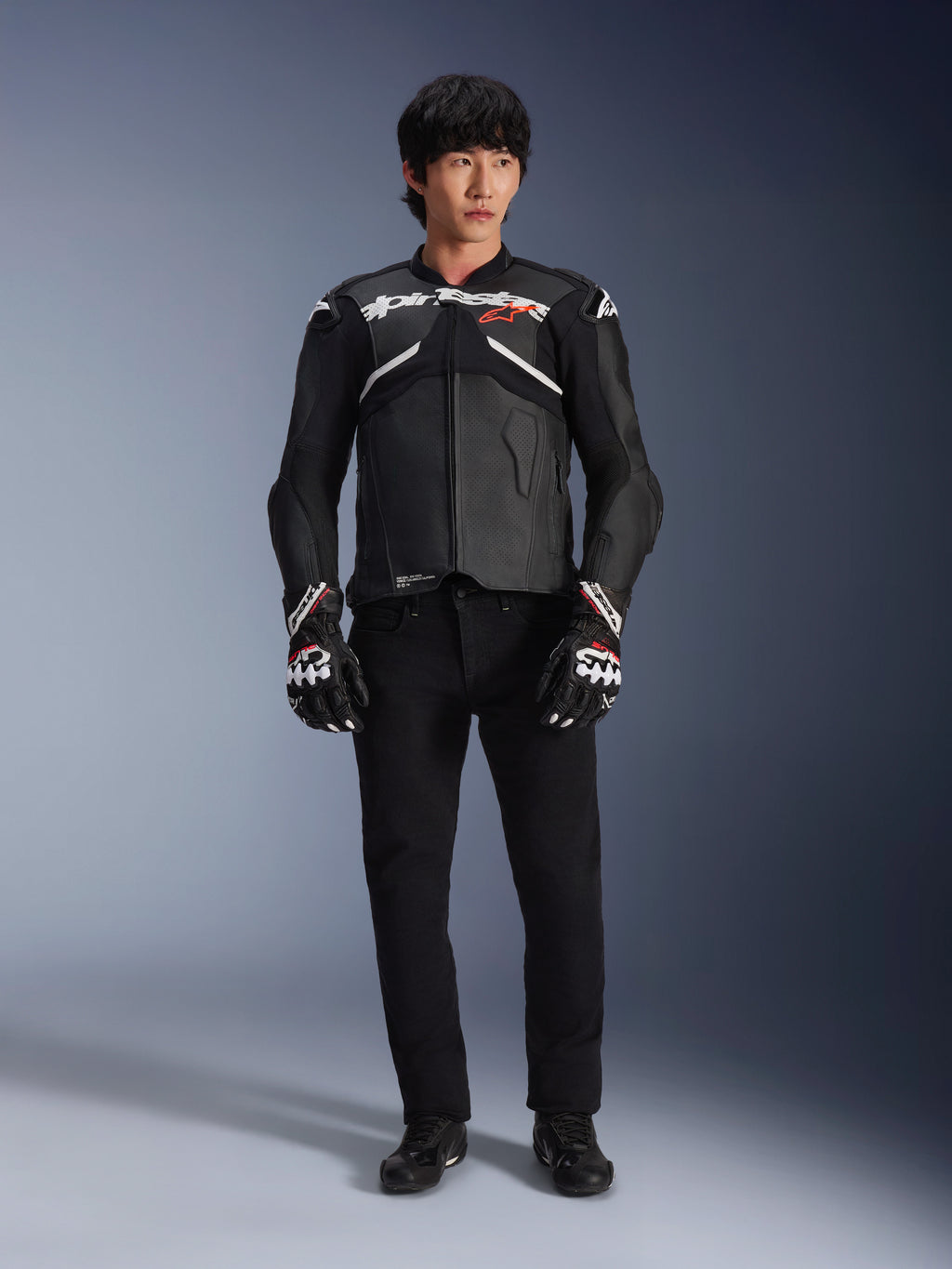 Modell som bär Alpinestars Atem V5 läderjacka i svart och vitt, med vit Alpinestars-logotyp och en röd stjärnlogga på bröstet, kombinerad med svarta byxor och svartvita racinghandskar, stilren motorcykelutrustning för hög prestanda