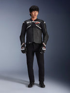 Modell som bär Alpinestars Atem V5 läderjacka i svart och vitt, med vit Alpinestars-logotyp och en röd stjärnlogga på bröstet, kombinerad med svarta byxor och svartvita racinghandskar, stilren motorcykelutrustning för hög prestanda