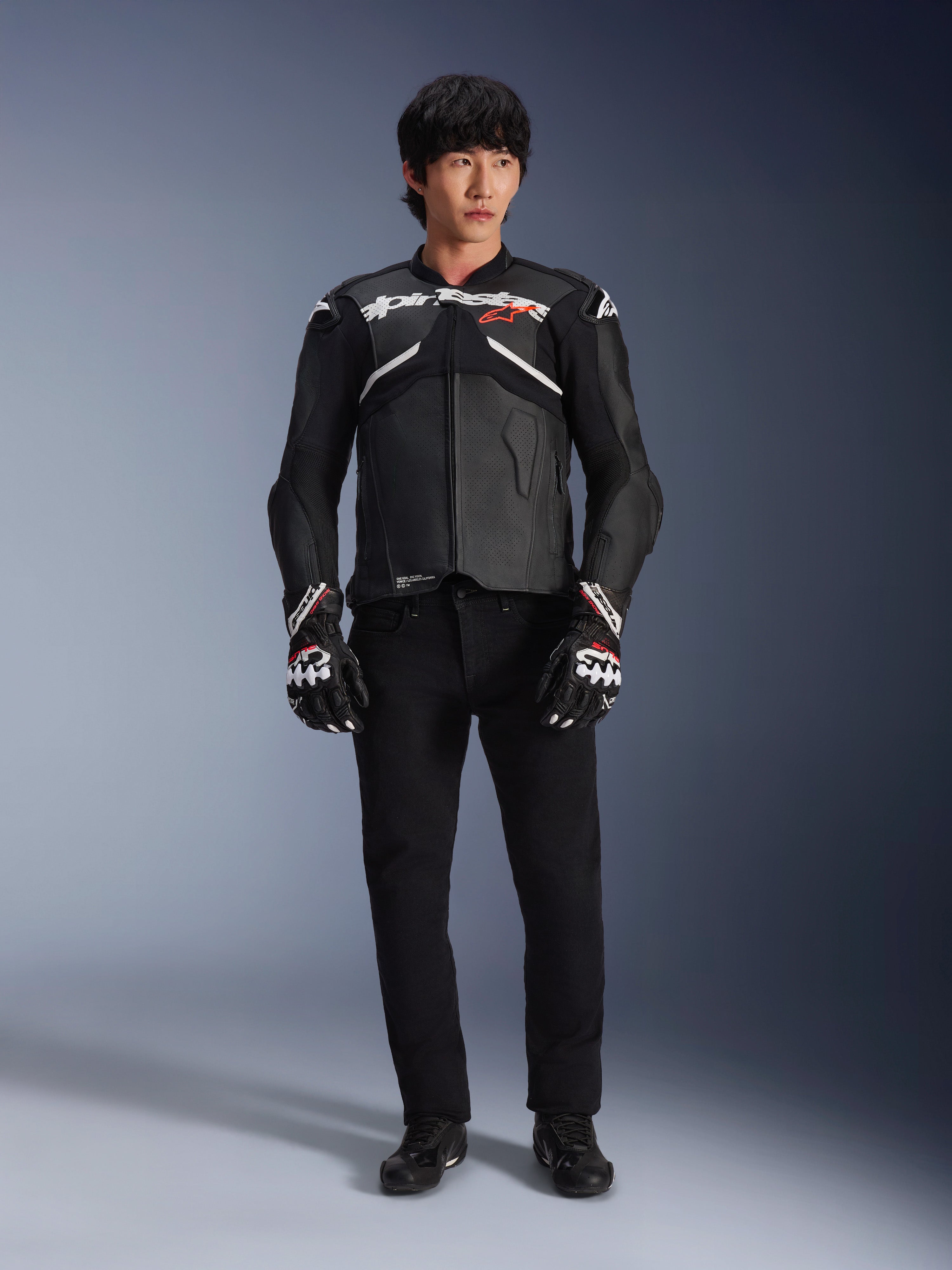 Modell som bär Alpinestars Atem V5 läderjacka i svart och vitt, med vit Alpinestars-logotyp och en röd stjärnlogga på bröstet, kombinerad med svarta byxor och svartvita racinghandskar, stilren motorcykelutrustning för hög prestanda