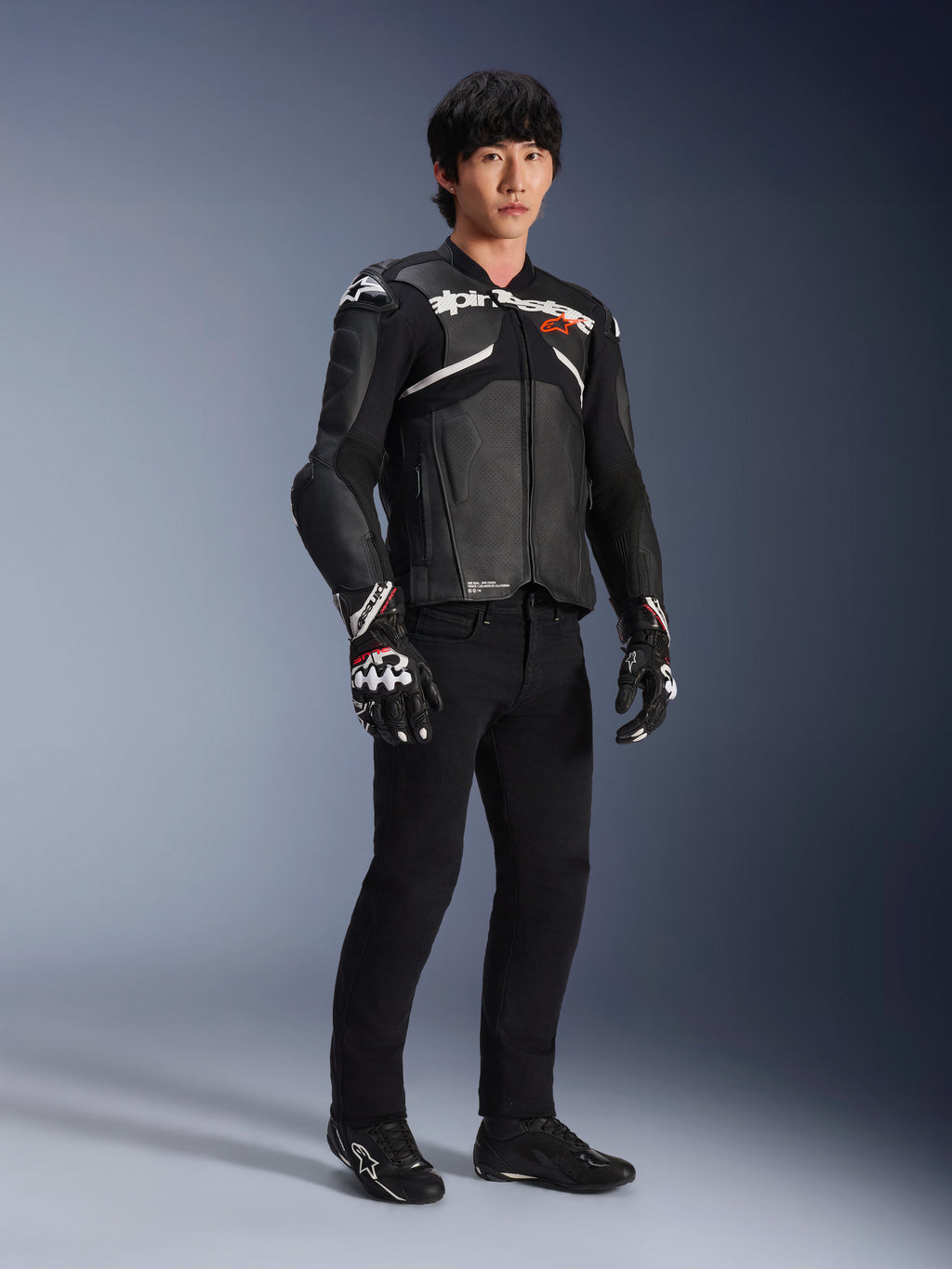 Alpinestars Atem V5 läderjacka, motorcykeljacka för racing, svart och vit, bärs med svarta byxor och matchande handskar, med vit Alpinestars-logotyp över bröstet och skyddande axelskydd.